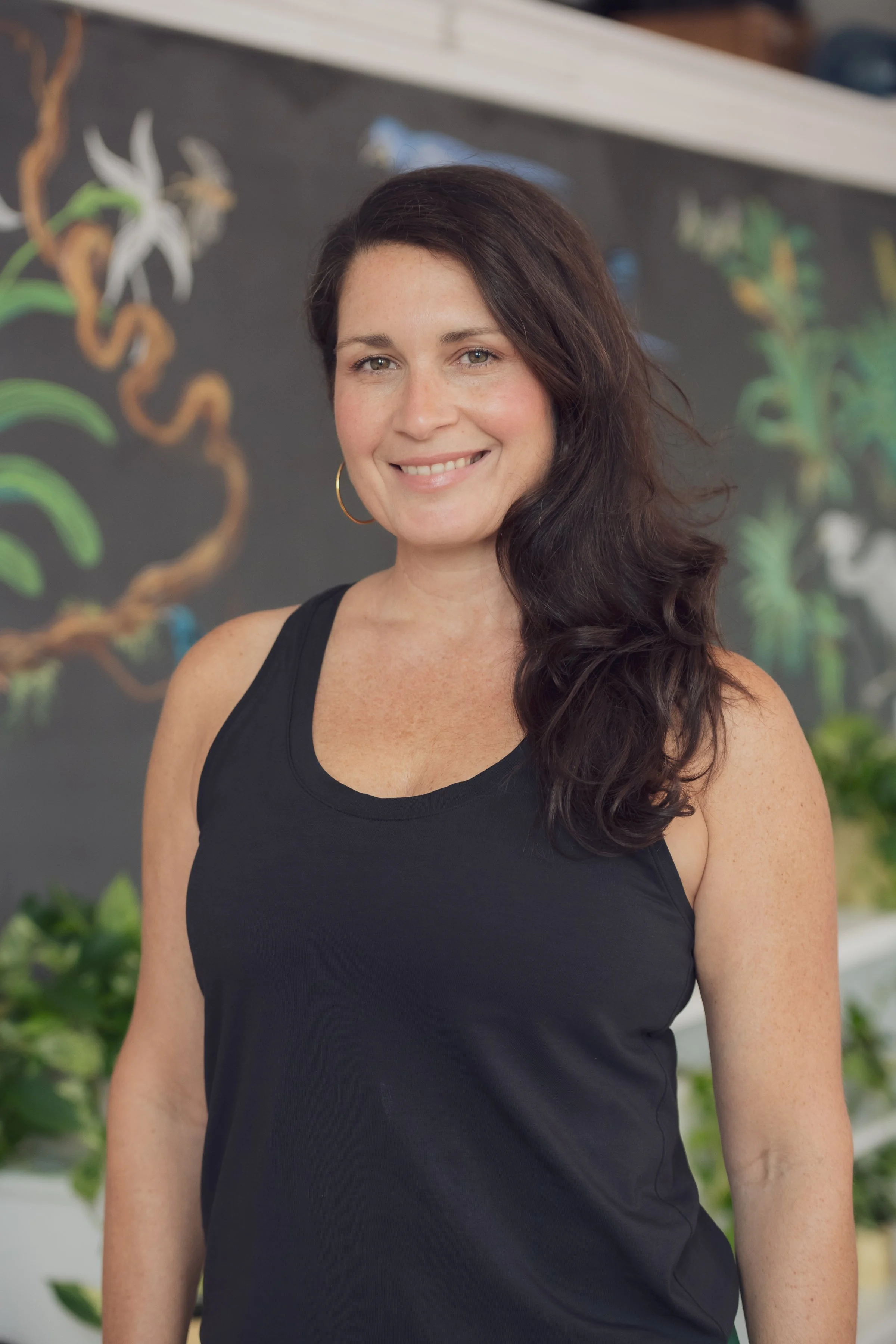 A smiling woman with long, dark hair, wearing a sleeveless black top, standing in front of a colorful mural painted on a wall.