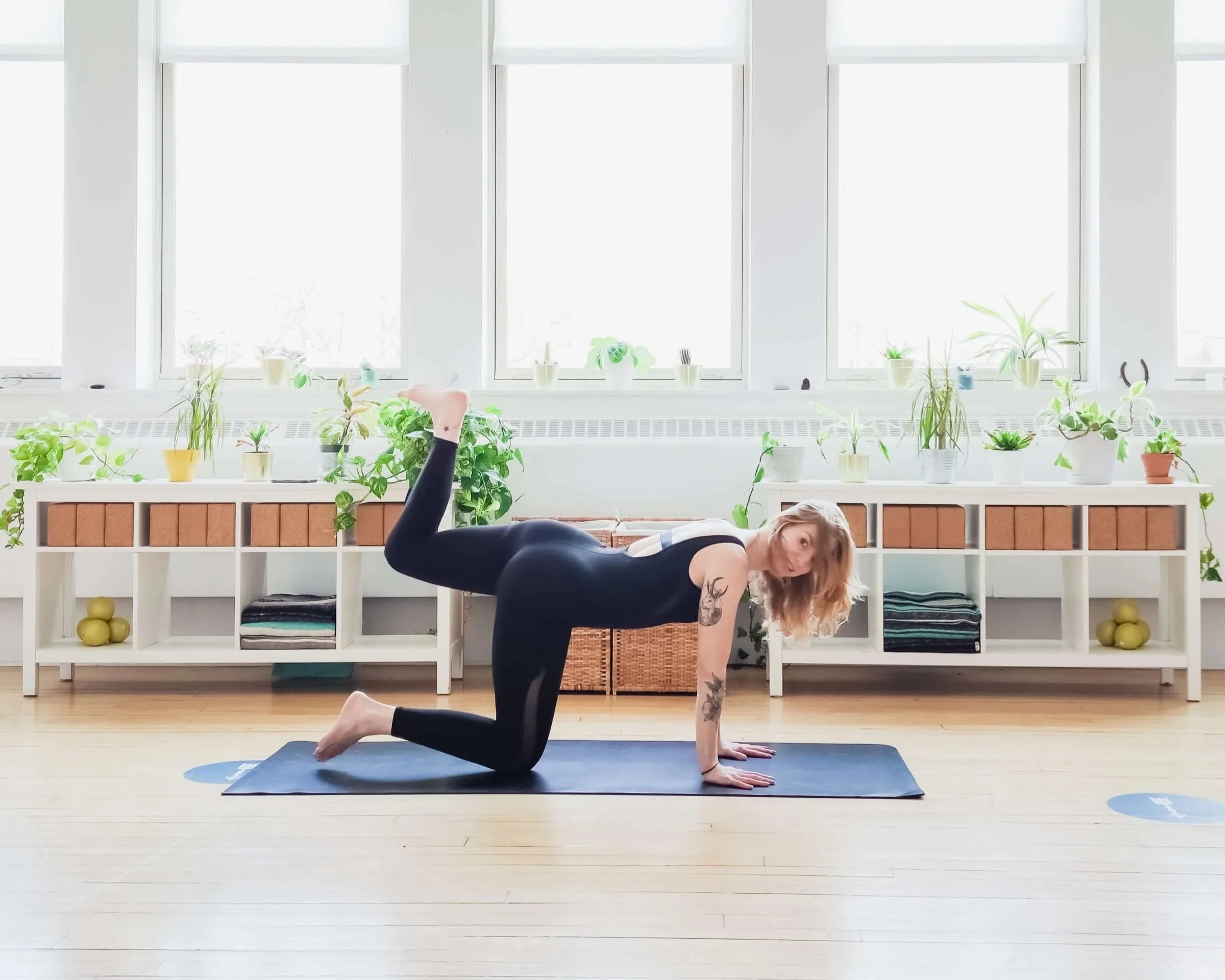 A woman with tattoos practicing yoga in a bright, plant-filled room, performing a three-legged downward dog pose on a yoga mat.