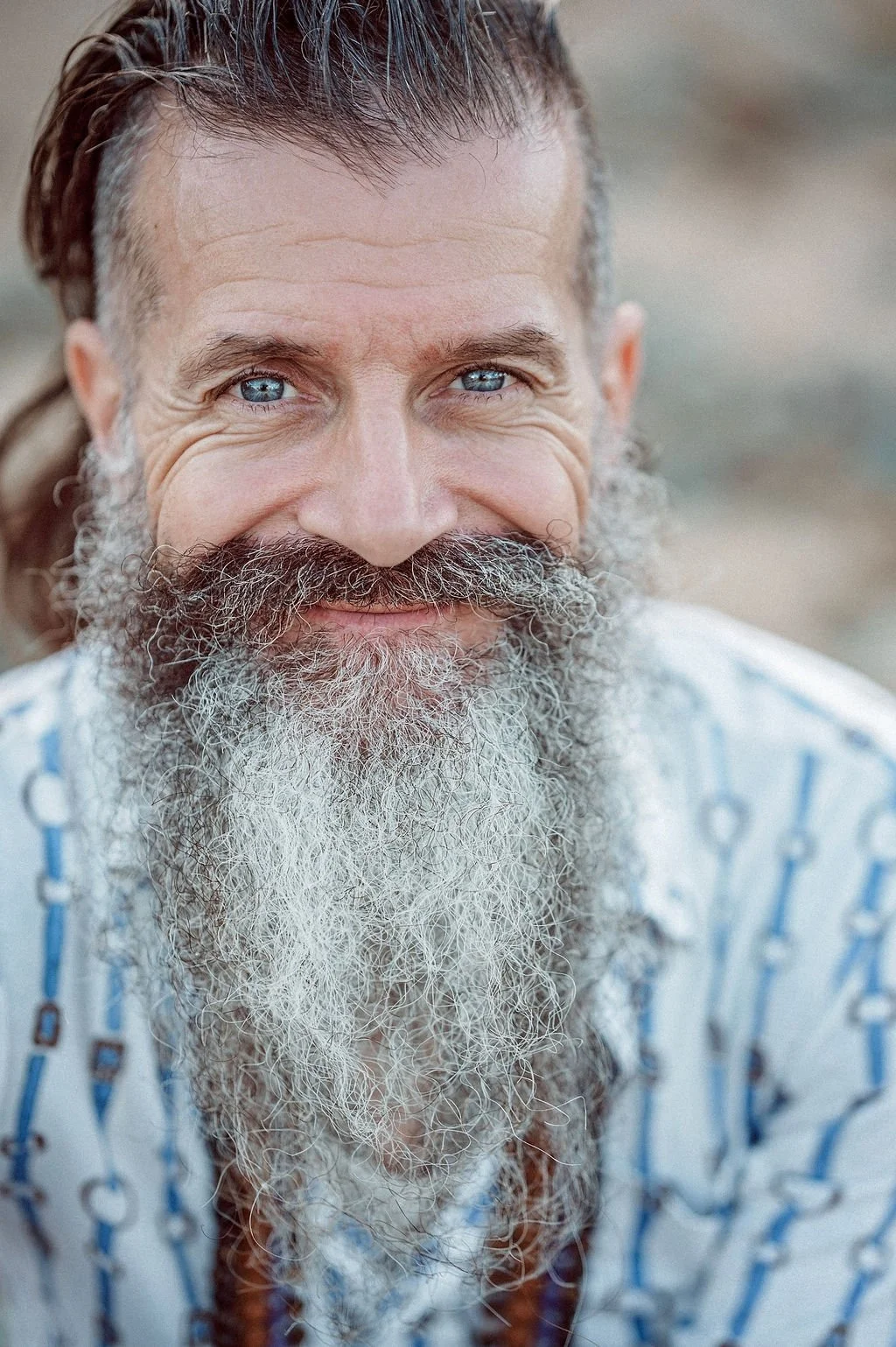 Close-up of a smiling man with a long gray and white beard, blue eyes, and slicked-back hair, wearing a patterned shirt.