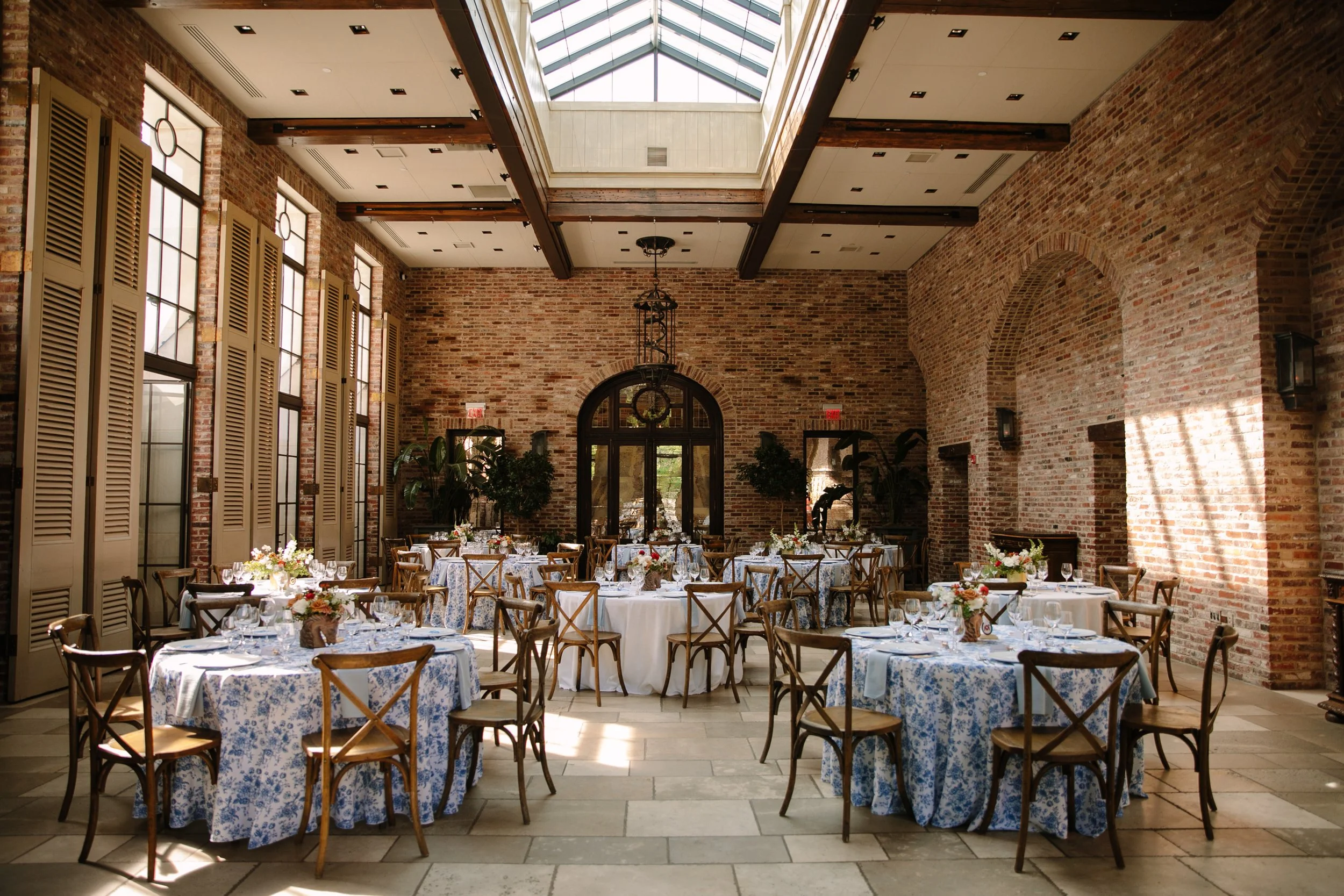 Indoor brick-walled banquet hall with round tables covered in blue and white floral tablecloths, set with glassware, silverware, and floral centerpieces, illuminated by natural light from a skylight.