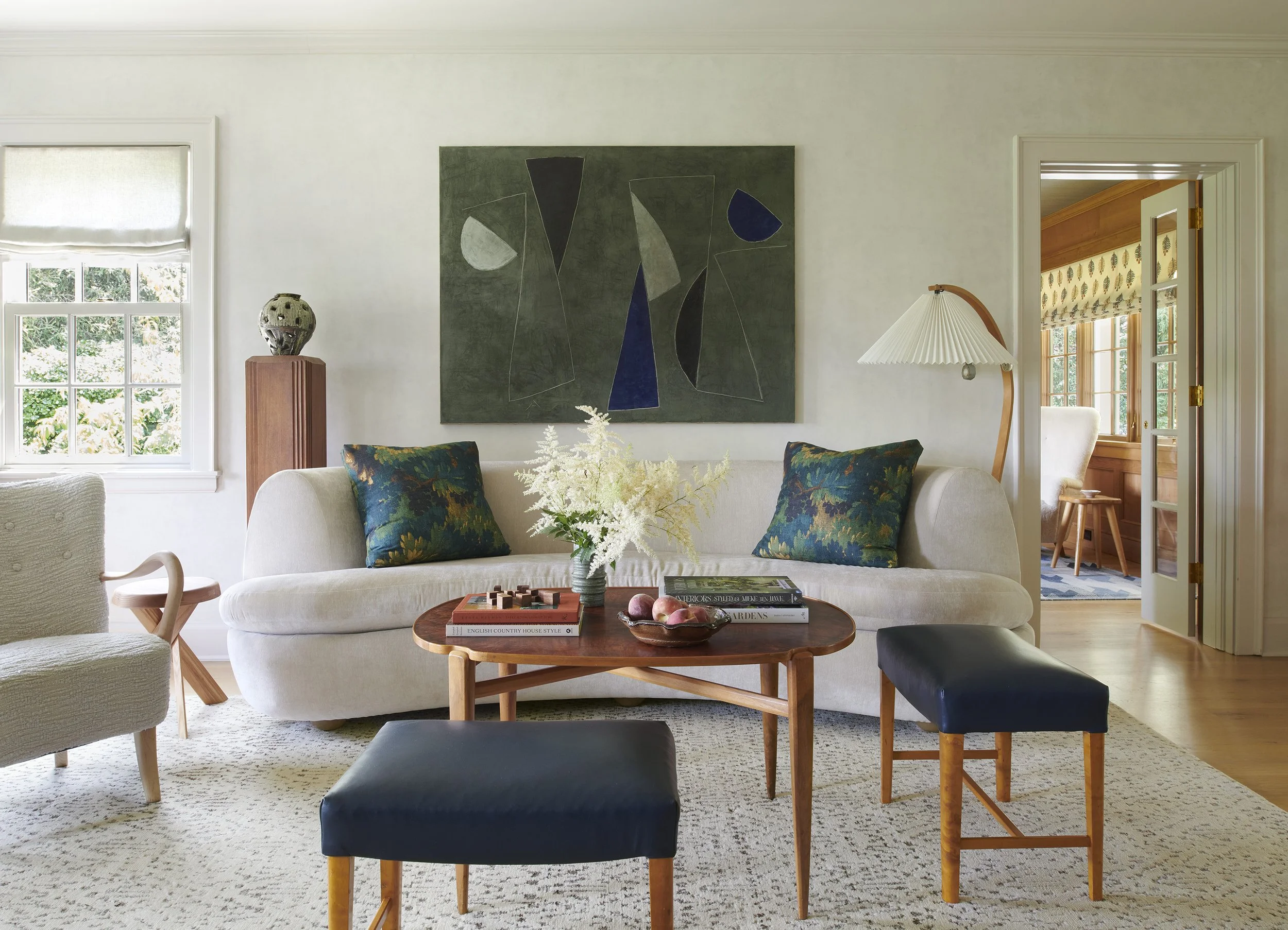 Elegant living room with a curved cream sofa, oval walnut coffee table, two navy leather ottomans, and a large dark green abstract painting above. Botanical print pillows, a sculptural armchair, and a pleated arc floor lamp complete the space.