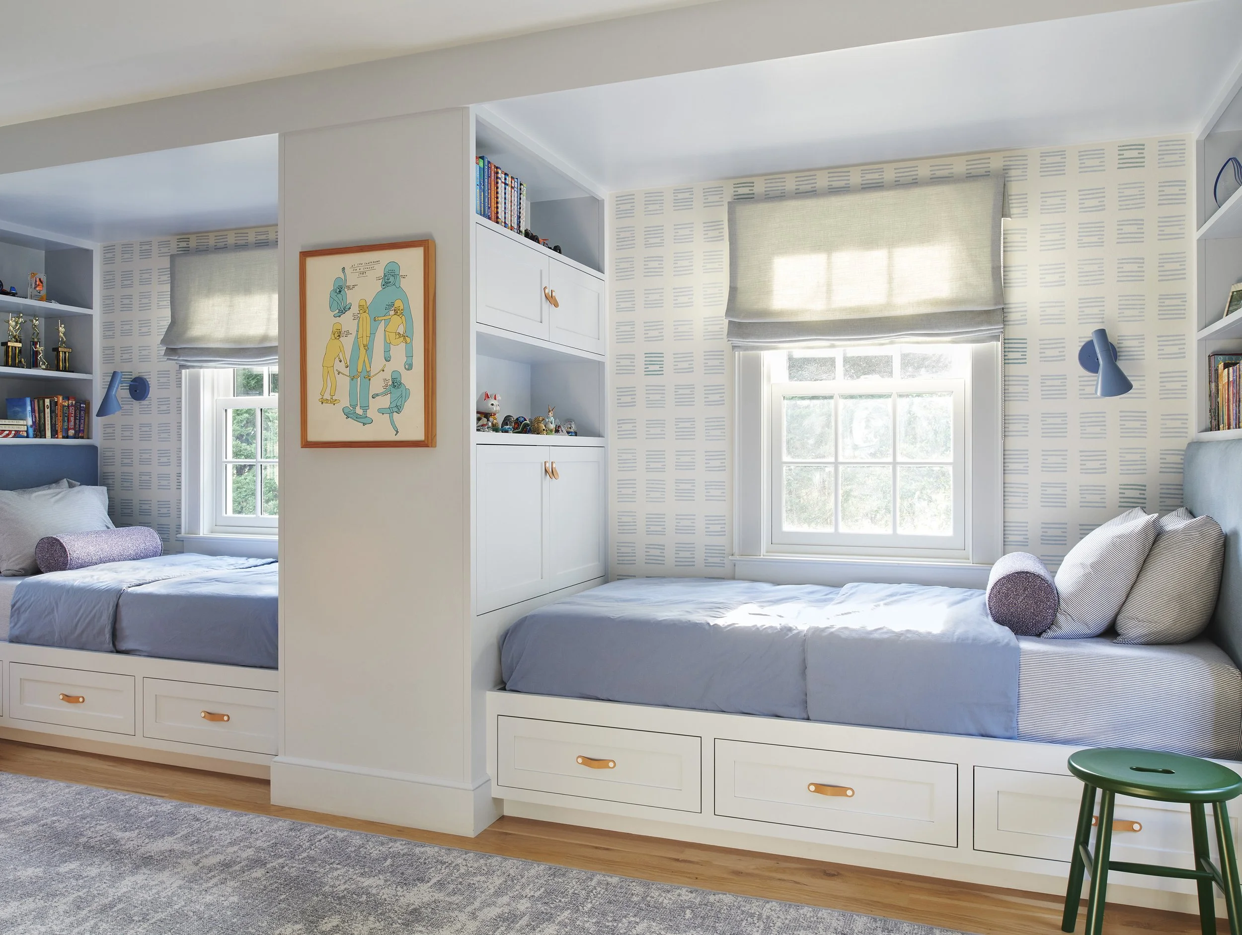 Shared children's bedroom with two built-in platform beds featuring under-bed storage drawers with leather pulls, blue bedding, and a central white built-in cabinet unit with open shelving.