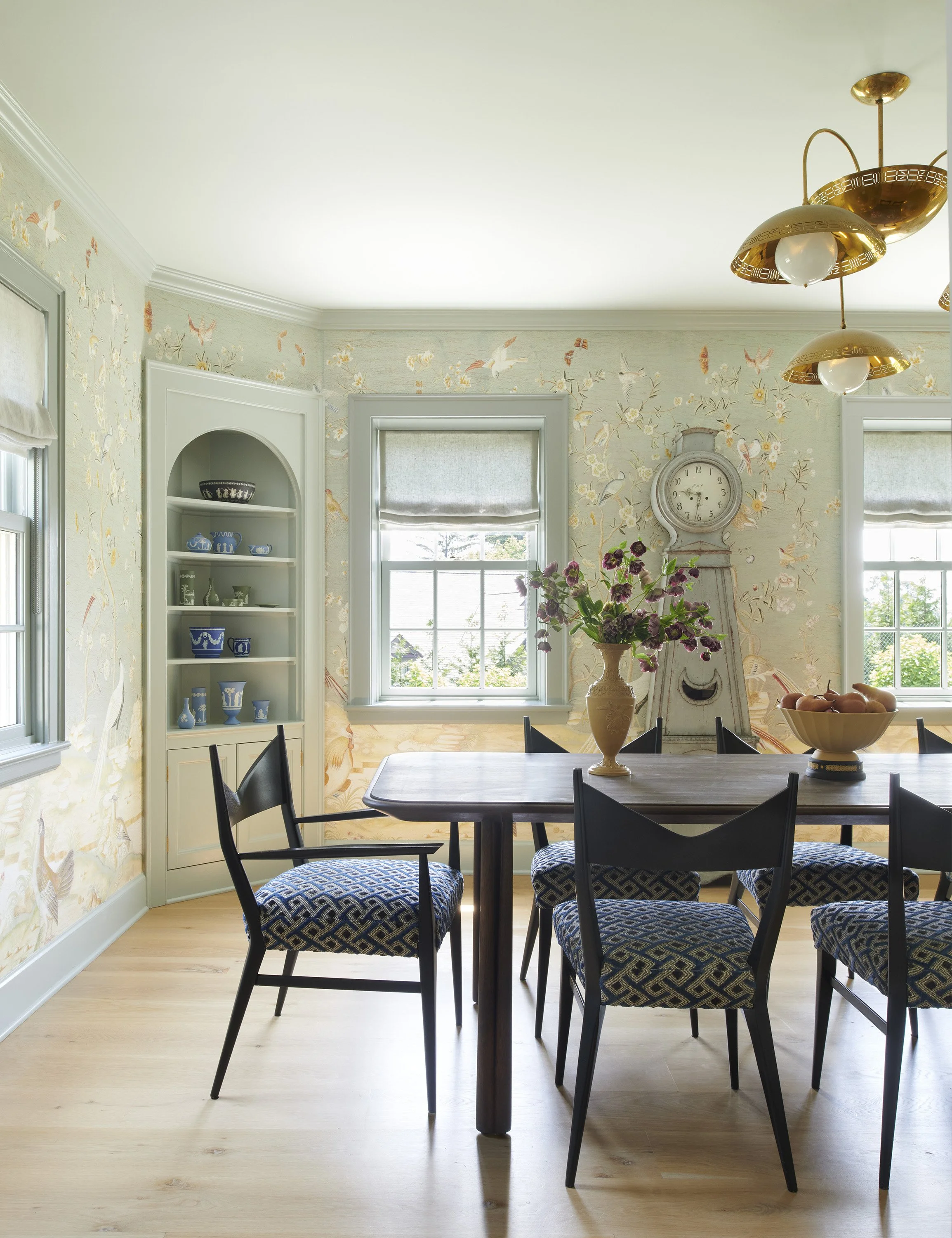 Dining room with floor-to-ceiling chinoiserie wallpaper featuring birds and botanicals, a dark wood dining table with blue geometric-upholstered chairs, a brass sputnik-style chandelier, built-in arched shelving displaying blue Wedgwood ceramics, and
