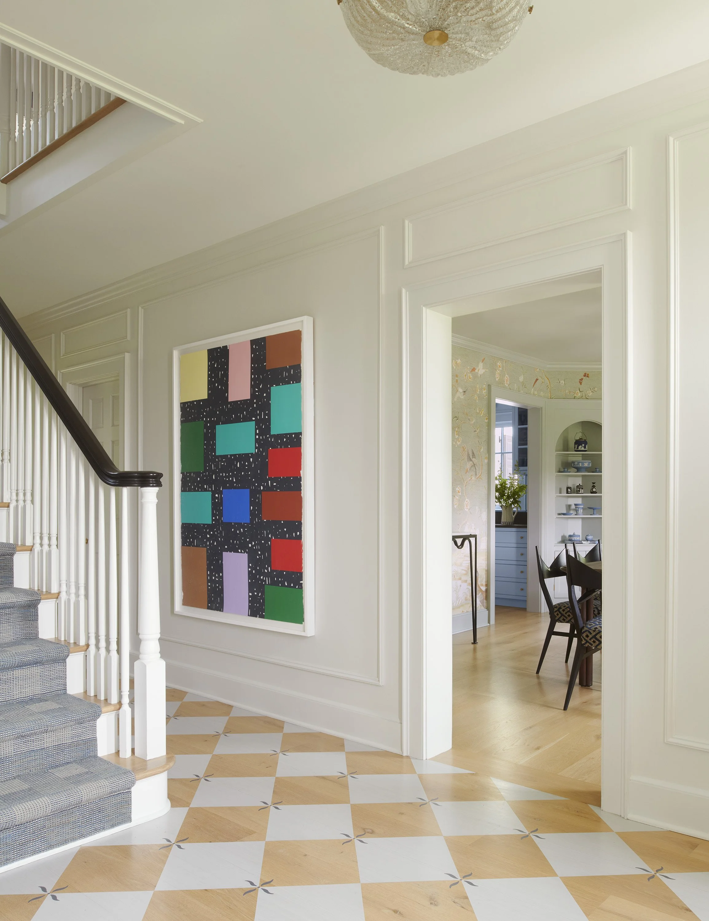 Bright entryway with a painted checkerboard wood floor, white paneled walls, and a large colorful grid-pattern abstract artwork. A staircase with dark wood banister and gray runner leads up on the left, with a glimpse into the chinoiserie dining room
