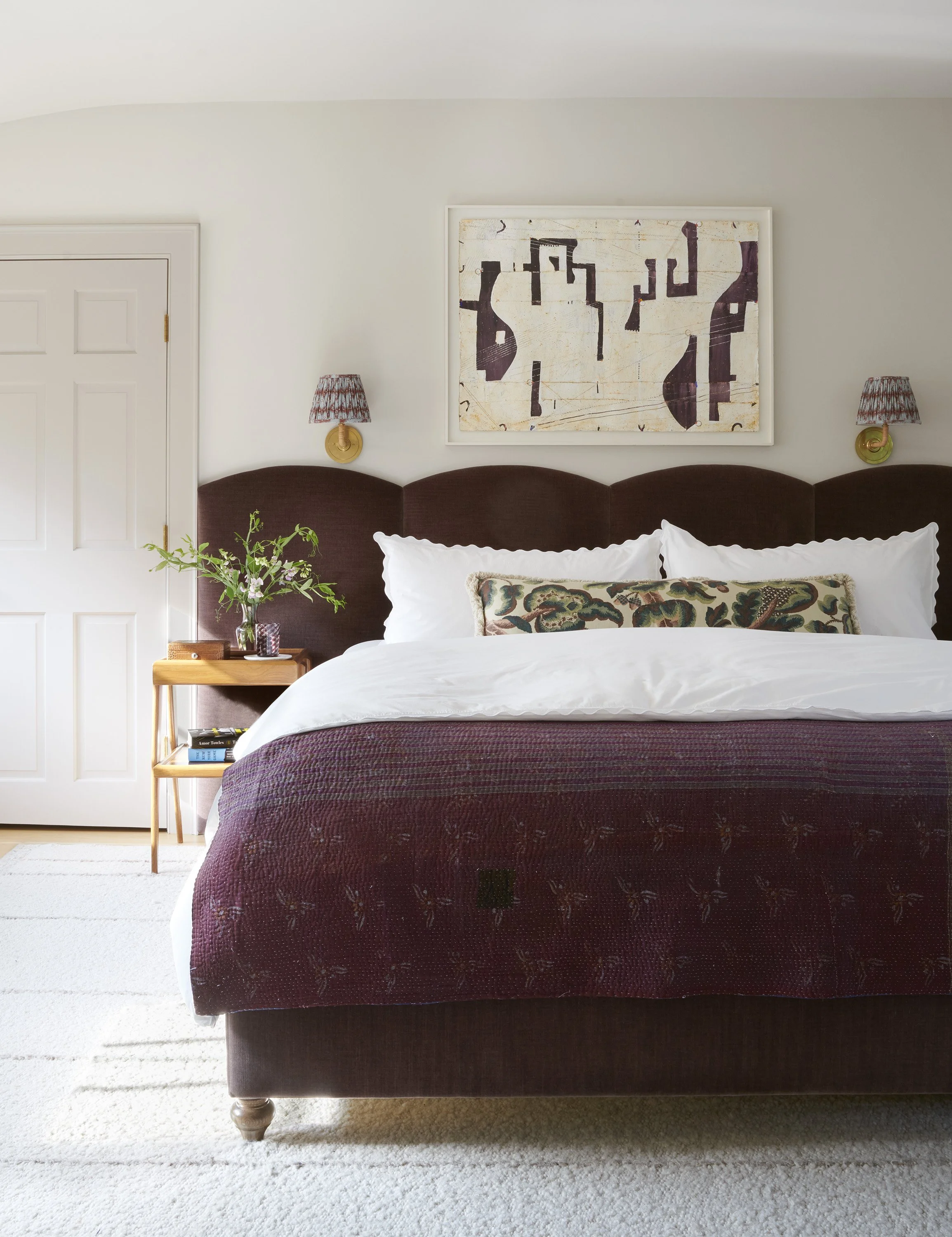 Bedroom featuring a deep burgundy upholstered bed with a scalloped headboard, white bedding, a vintage kantha quilt, and a floral lumbar pillow. A framed abstract artwork hangs above flanked by brass wall sconces with printed shades. A light wood nig
