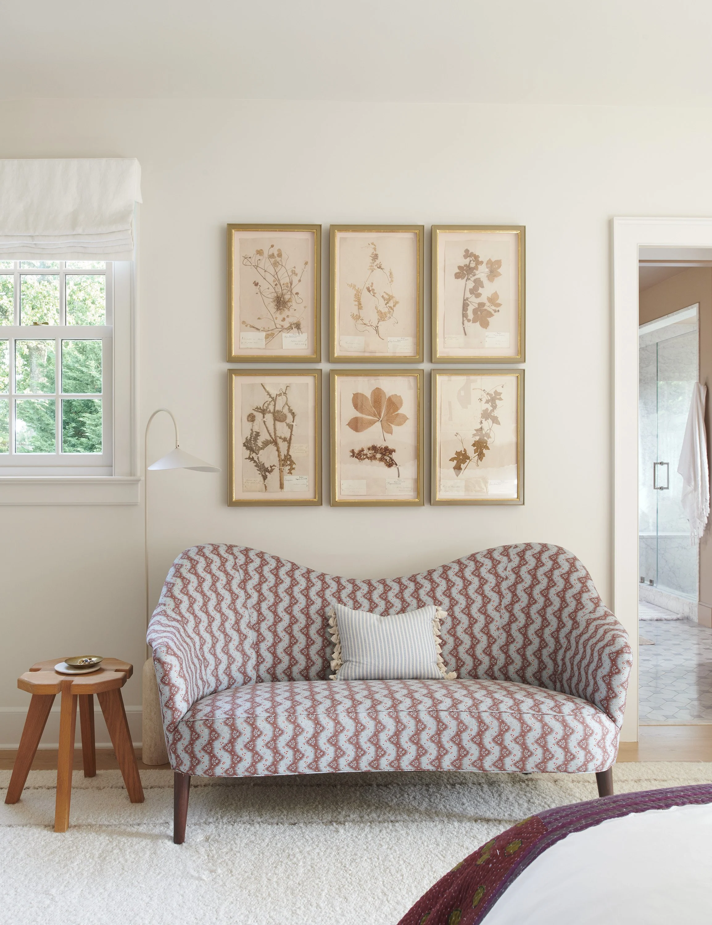 An elegant, sun-drenched reading nook with a patterned camelback sofa, six gold-framed pressed leaf illustrations, a simple wooden stool, and a soft white plush rug.