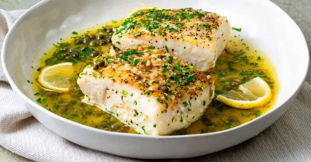Modern wooden house with large windows beside a bush, surrounded by tall grass and mountains in the background.