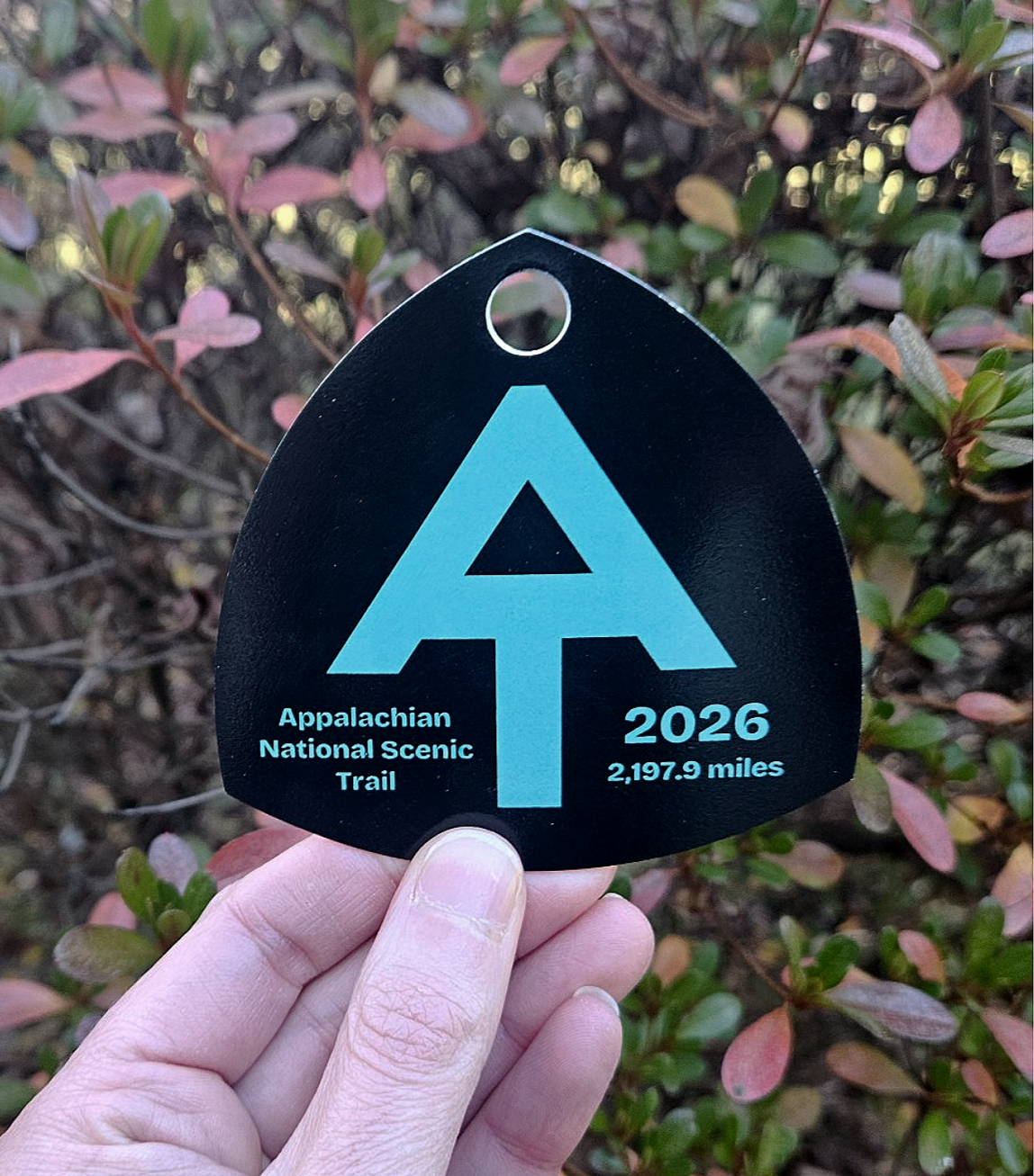 Appalachian Trail hiker registration hang tag with the A.T. logo and mileage, held in front of foliage.