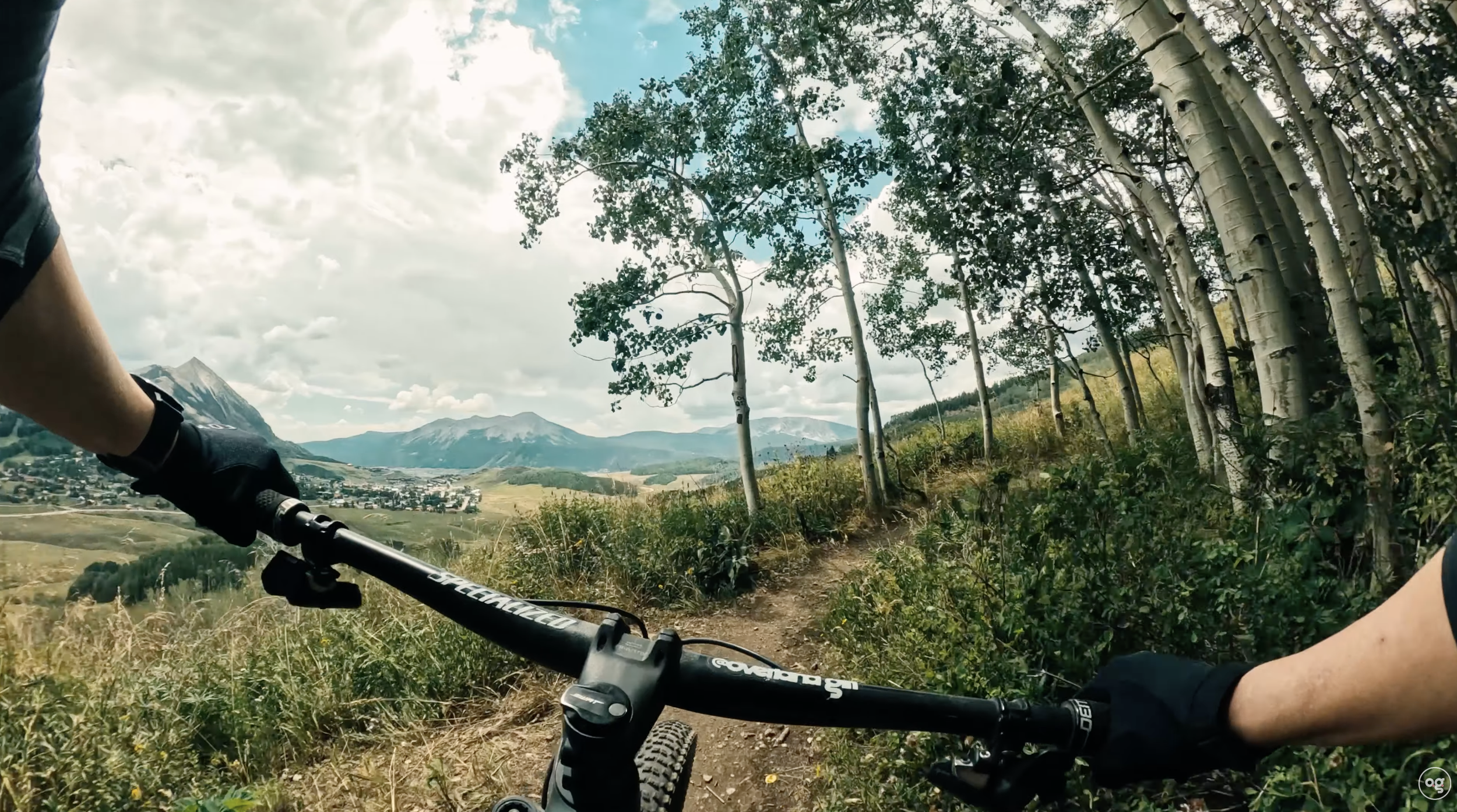 First-person mountain biking view on Snodgrass Trail, with singletrack winding past aspen trees and Mt. Crested Butte in the background, Colorado.