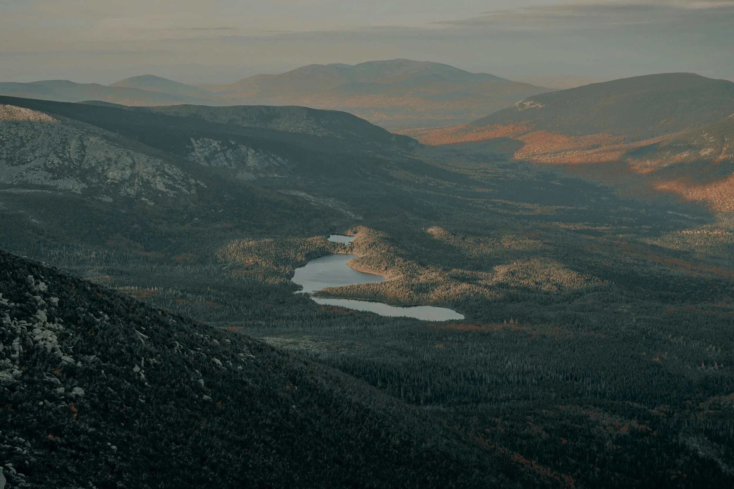 High-angle view of forested mountains and connected lakes in northern Maine with evening light across the landscape.
