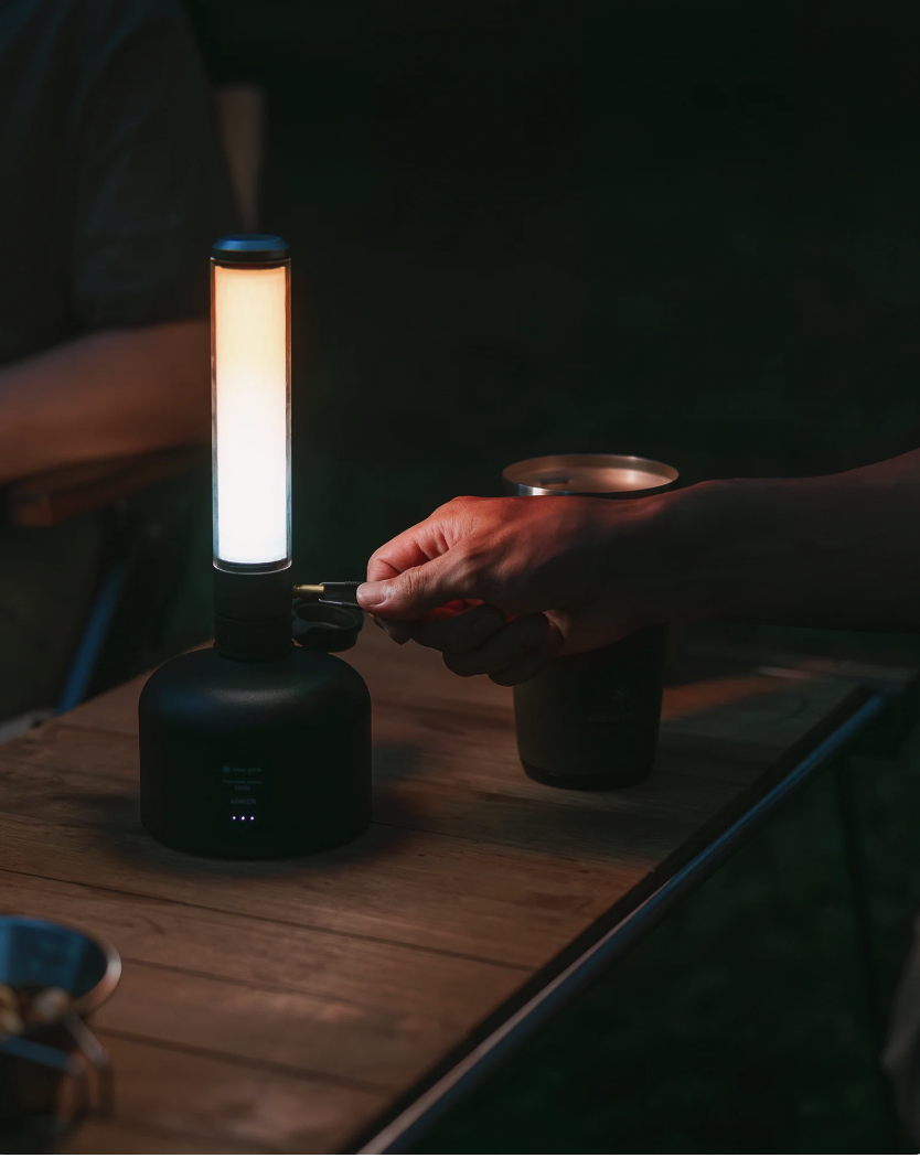 A hand adjusts the flame on a Snow Peak GigaPower Tabletop Lantern on a wooden camp table at night.