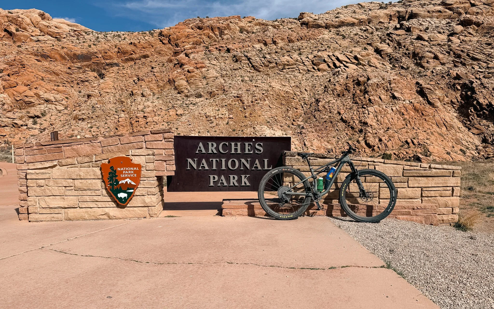 : Specialized mountain bike parked in front of the Arches National Park entrance sign near Moab, Utah, with red sandstone cliffs in the background.