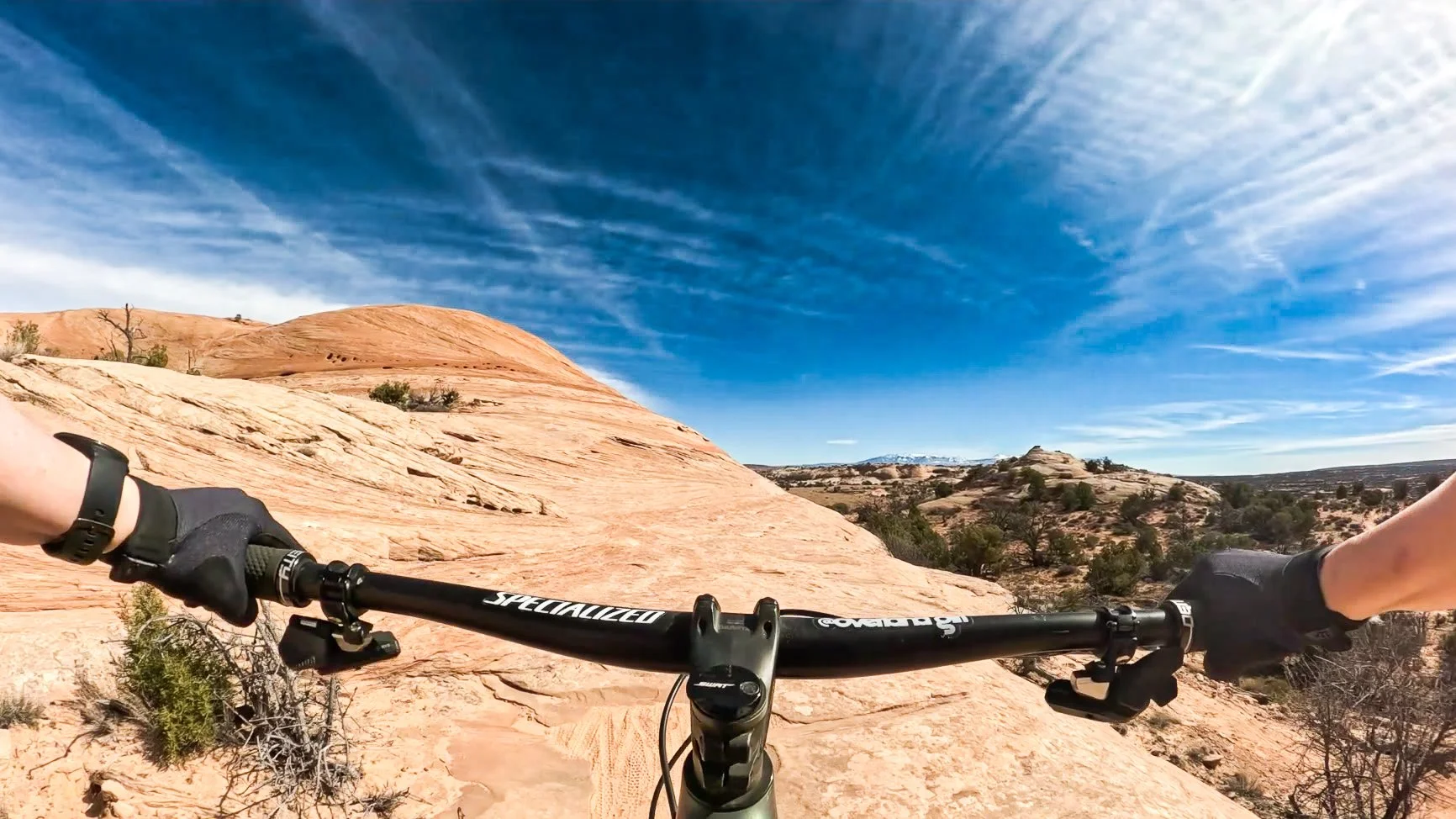 First-person view mountain biking Ramblin' Trail at Navajo Rocks near Moab, Utah, riding a Specialized mountain bike across sandstone slickrock with desert mesas and a blue sky.