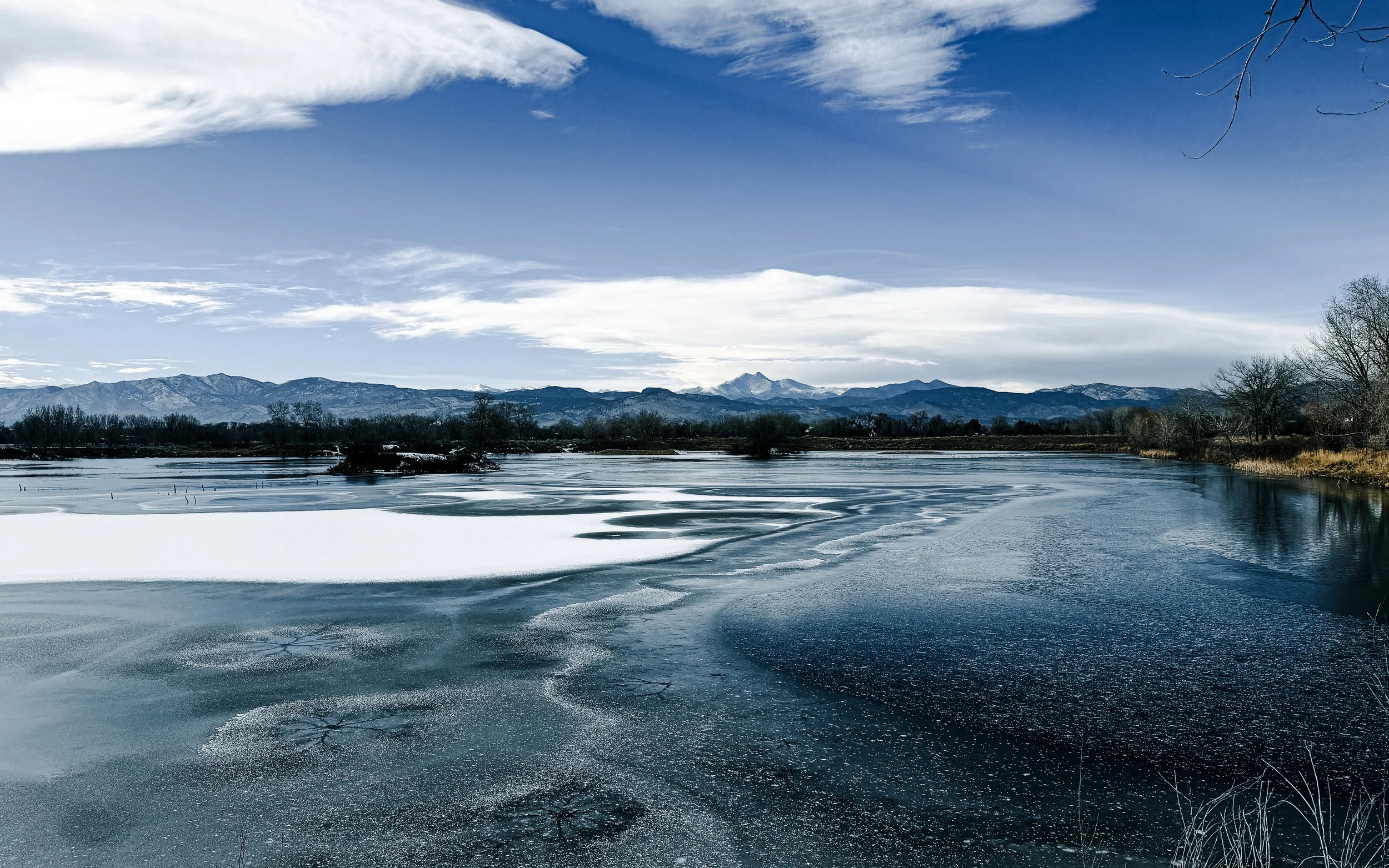 Partially iced Heron Lake at Pella Crossing Park near Hygiene, Colorado with the Rocky Mountains in the distance under winter skies.