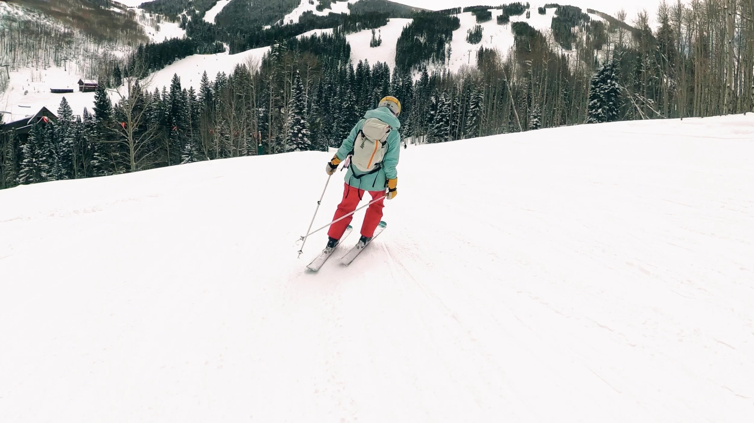Skier in a mint green jacket and red pants descending a snowy run at Beaver Creek, with pine trees, mountain lift lines, and ski village buildings visible in the background.
