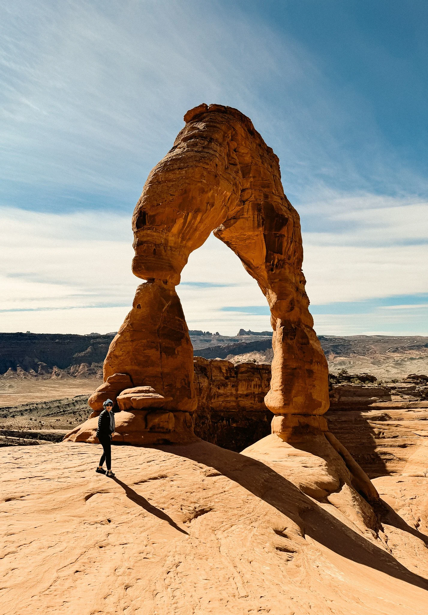 Person walking toward Delicate Arch in Arches National Park with massive sandstone arch towering above desert landscape