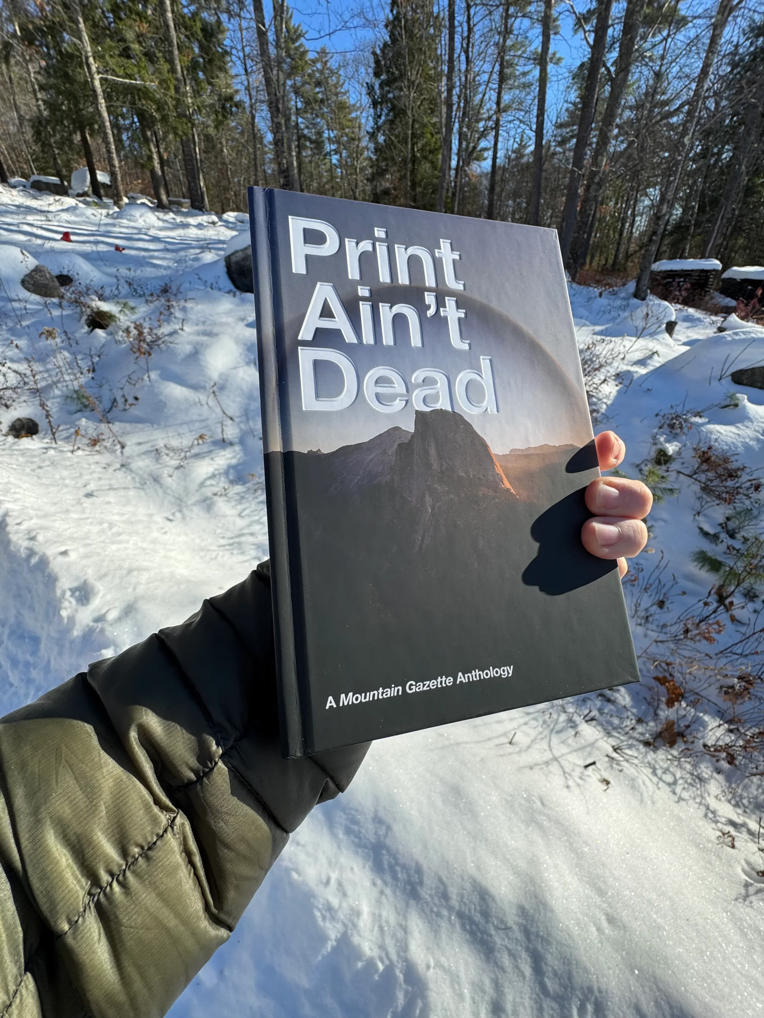 Hand holding Mountain Gazette's Print Ain't Dead anthology in a snowy forest landscape.