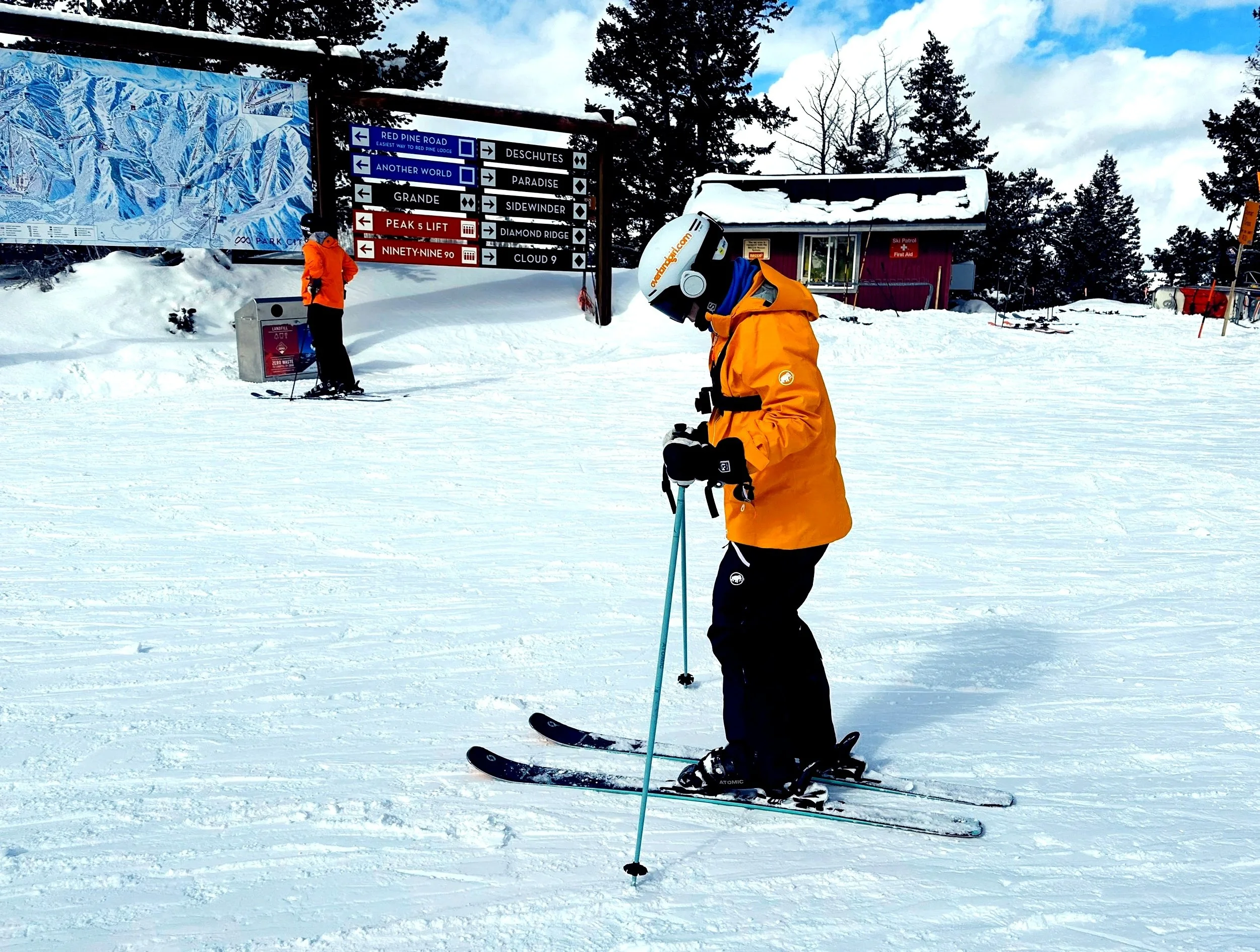 Skier in an orange Mammut shell jacket wearing Smartwool Classic Thermal Merino base layers underneath, standing at the top of a run at Park City ski resort with trail signs and a snow-covered patrol building in the background.