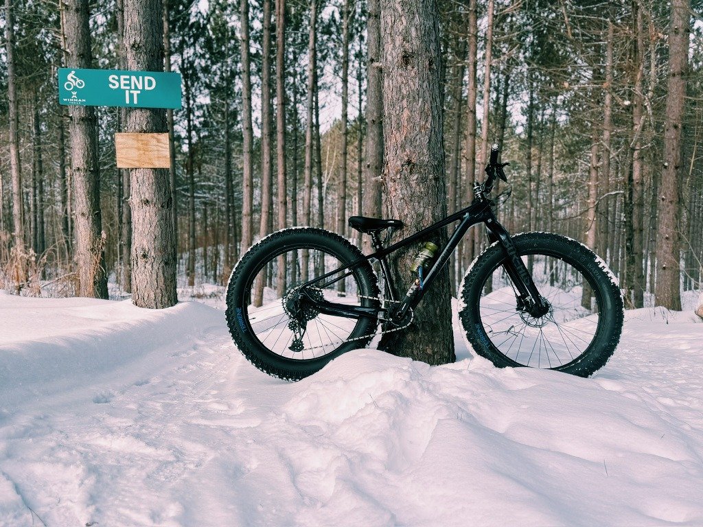A Borealis Crestone fat bike resting against a tree at Winman Trails in Northern Wisconsin, positioned at the top of the Send It trail on a snowy day.