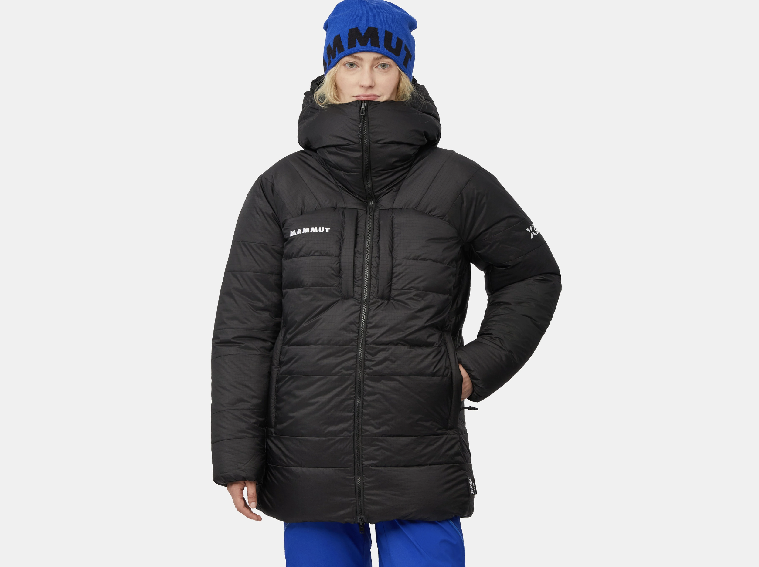Front view of a woman wearing a black Mammut Eiger Nordwand Pro Down IN Parka with an insulated hood and blue Mammut beanie, hands in pockets, on a light gray studio background.