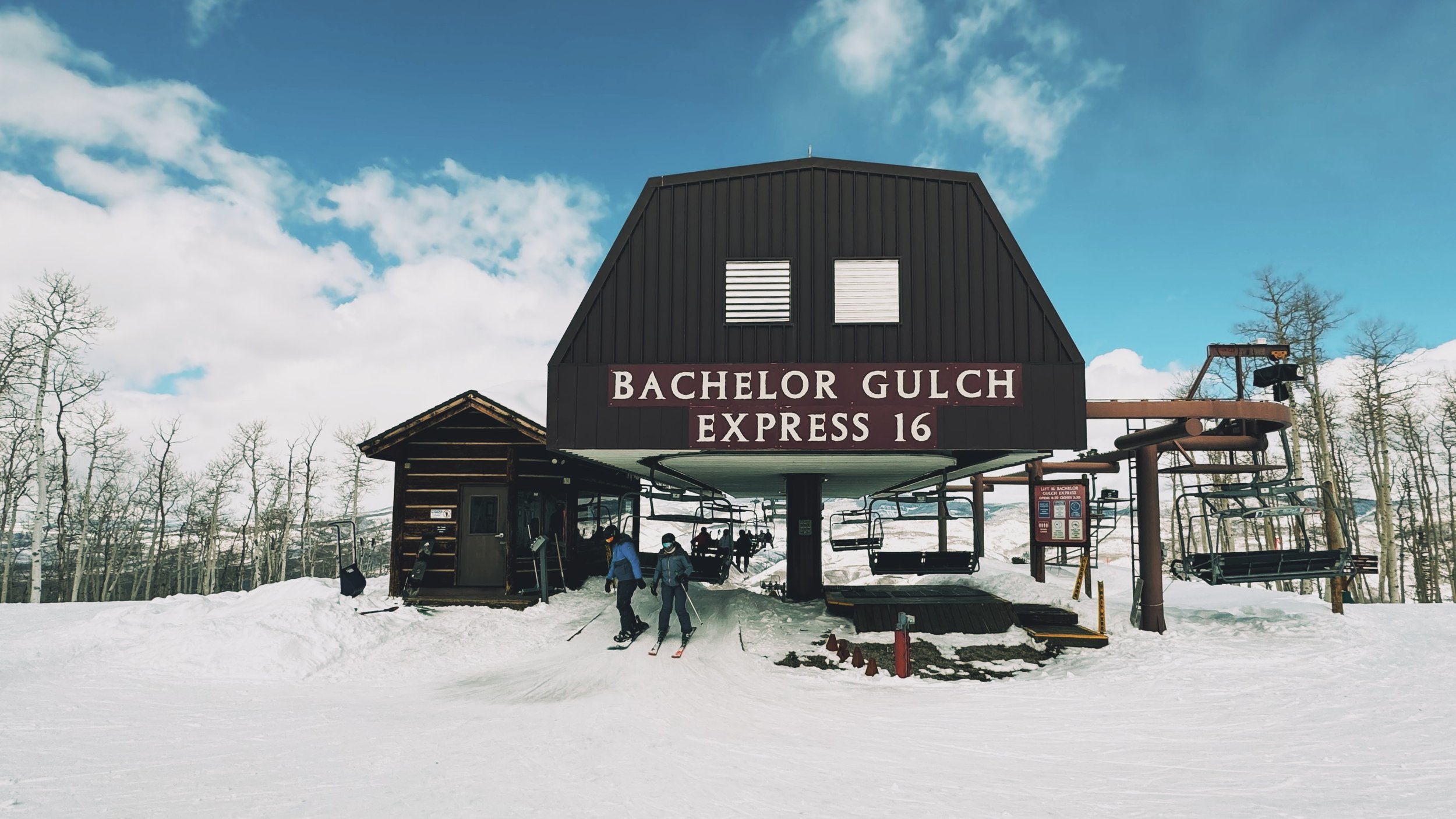 Skiers and snowboarders getting off Bachelor Gulch Express Lift (#16) at the top, with a scenic mountain view in the background.