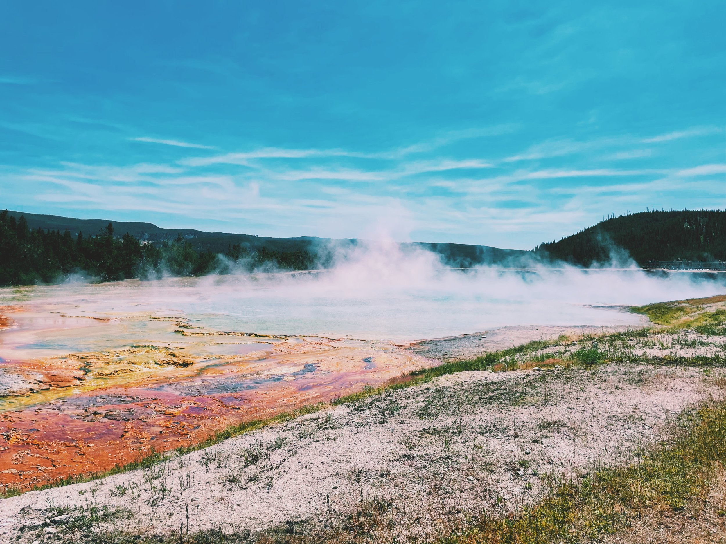 Excelsior Geyser Crater with hot water flowing into the Firehole River, surrounded by steam and rocky landscape.
