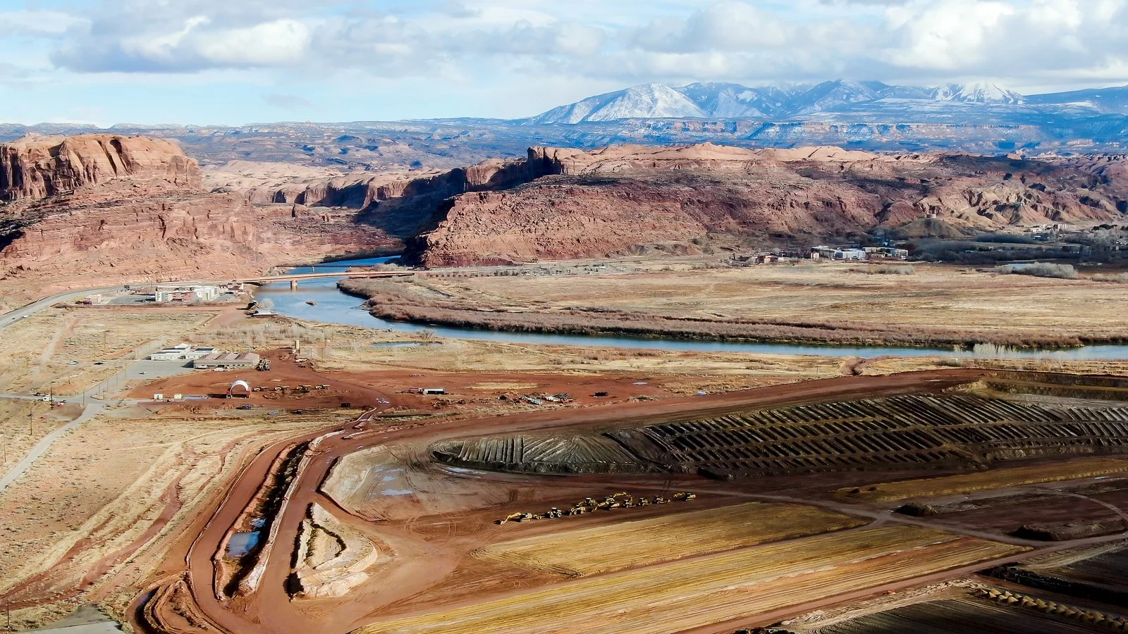 Aerial view of the former Atlas uranium mill tailings site near Moab, Utah, showing earthmoving equipment and layered waste piles beside the Colorado River with red rock canyon walls in the background.