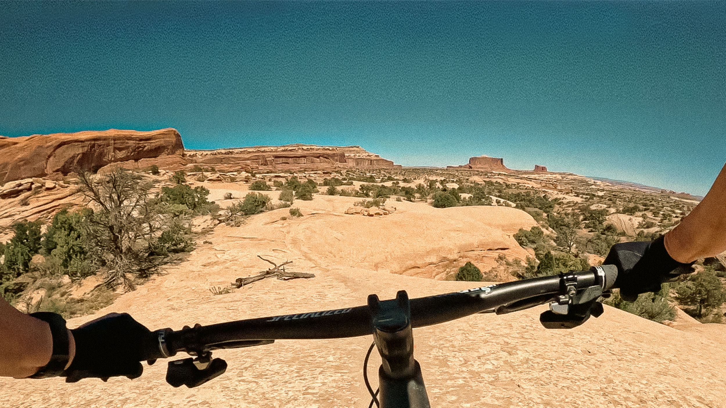 First-person view mountain biking Ramblin' Trail at Navajo Rocks near Moab, Utah, with a Specialized mountain bike on sandstone slickrock overlooking desert mesas and red rock formations.