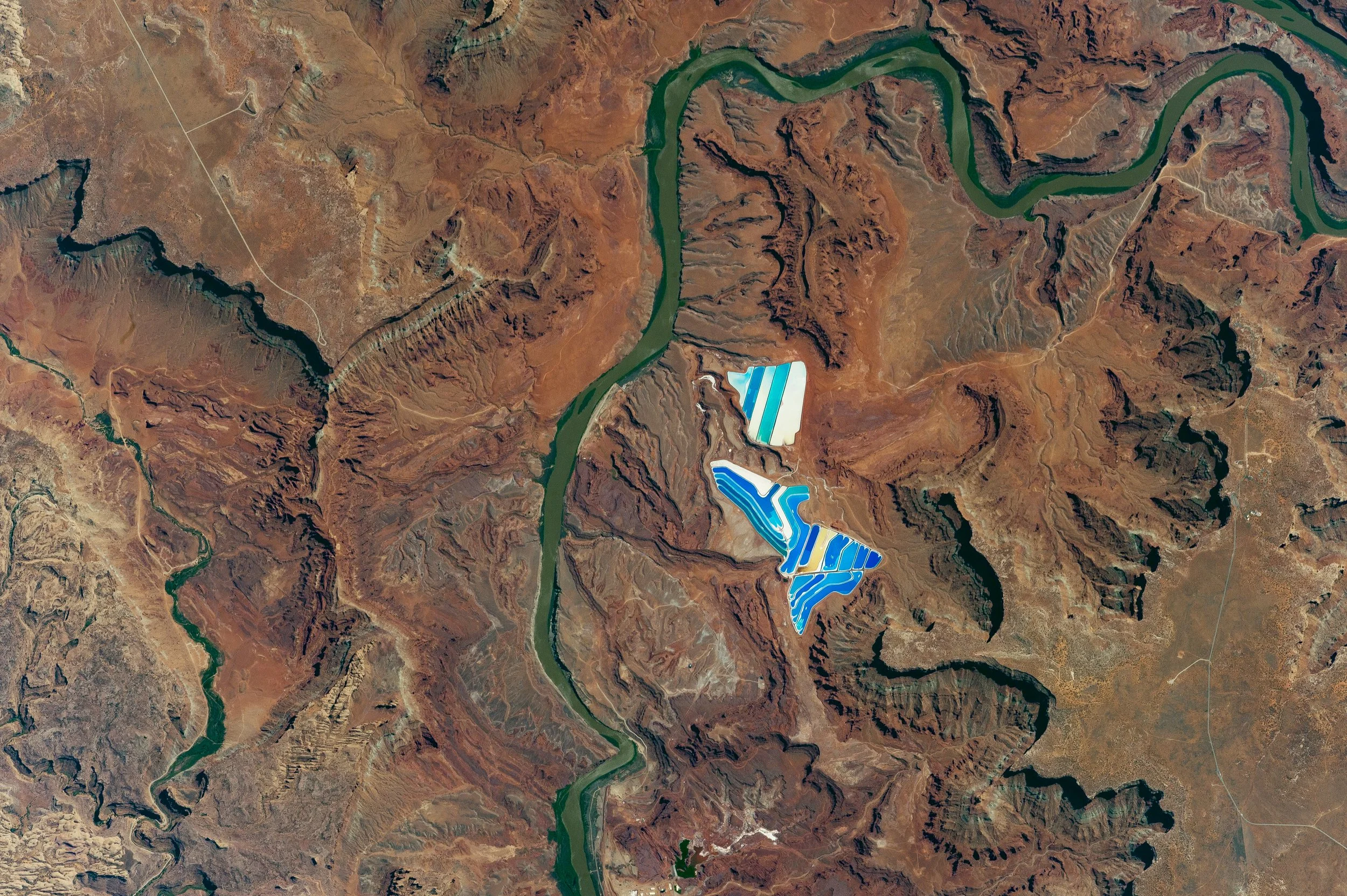 Satellite view of the Colorado River winding through canyon country near Moab, with blue and white potash evaporation ponds visible against red rock desert.