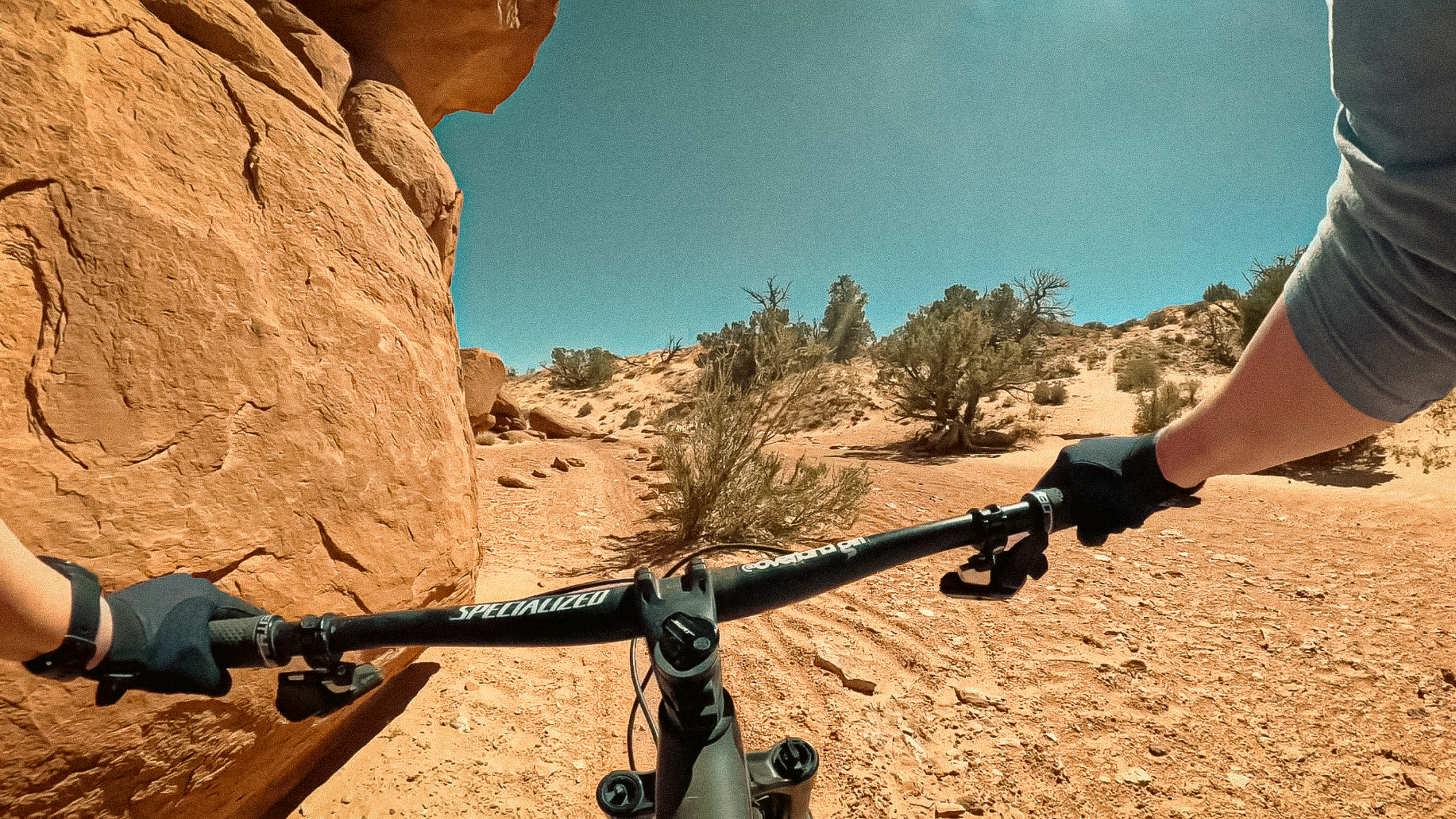 First-person view mountain biking Big Mesa Trail at Navajo Rocks near Moab, Utah, riding a Specialized mountain bike past a large sandstone rock formation on a rocky desert trail.