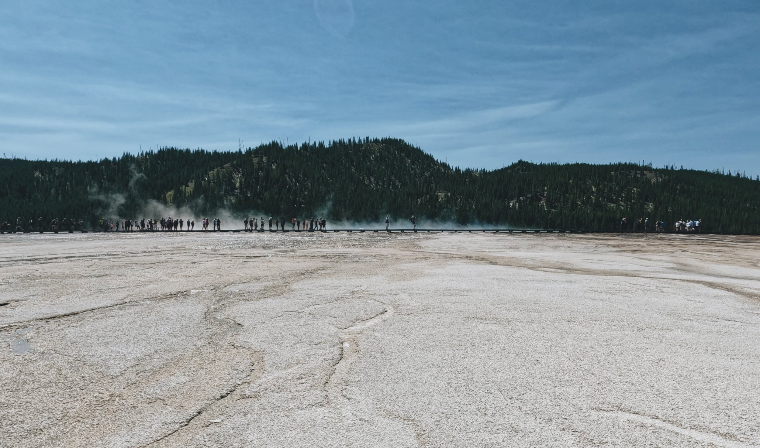 Visitors walk on elevated boardwalks through Midway Geyser Basin, surrounded by steam and colorful hot springs.