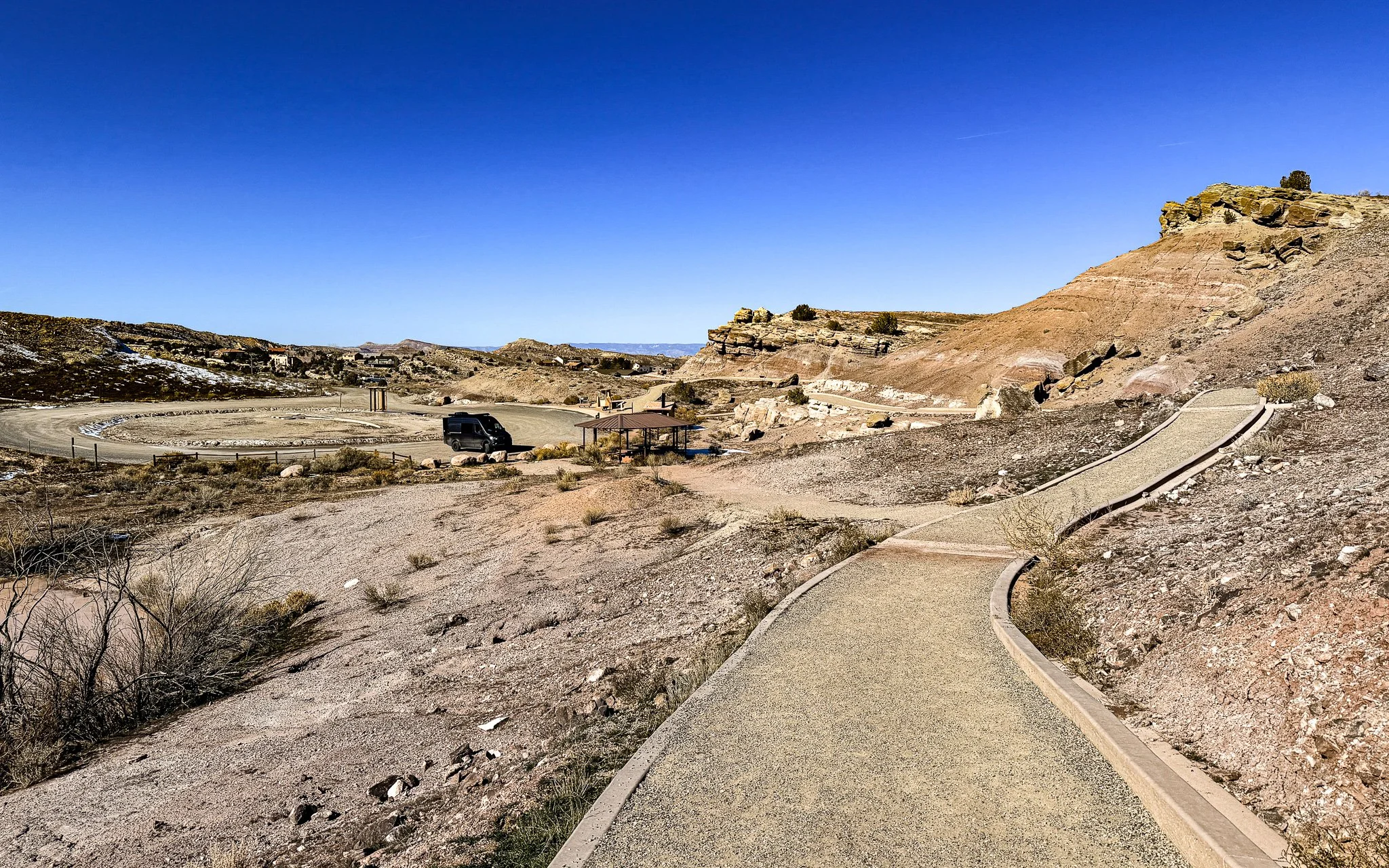 Concrete walking path winding through rocky desert hills at Dinosaur Hill in Fruita, Colorado, with fossil-bearing slopes and a trailhead area in the distance.