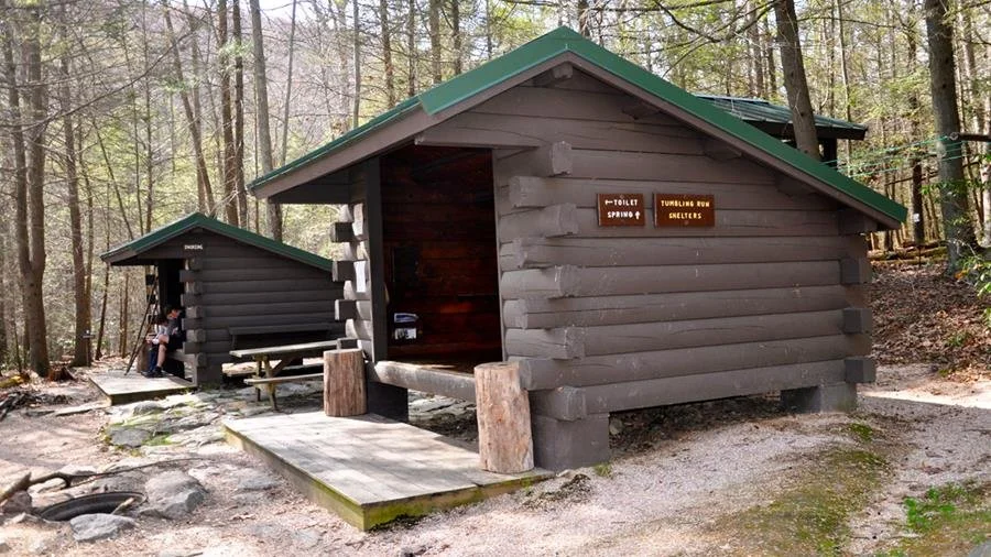 Two Appalachian Trail shelters with wooden platforms and signage in a wooded campsite area.