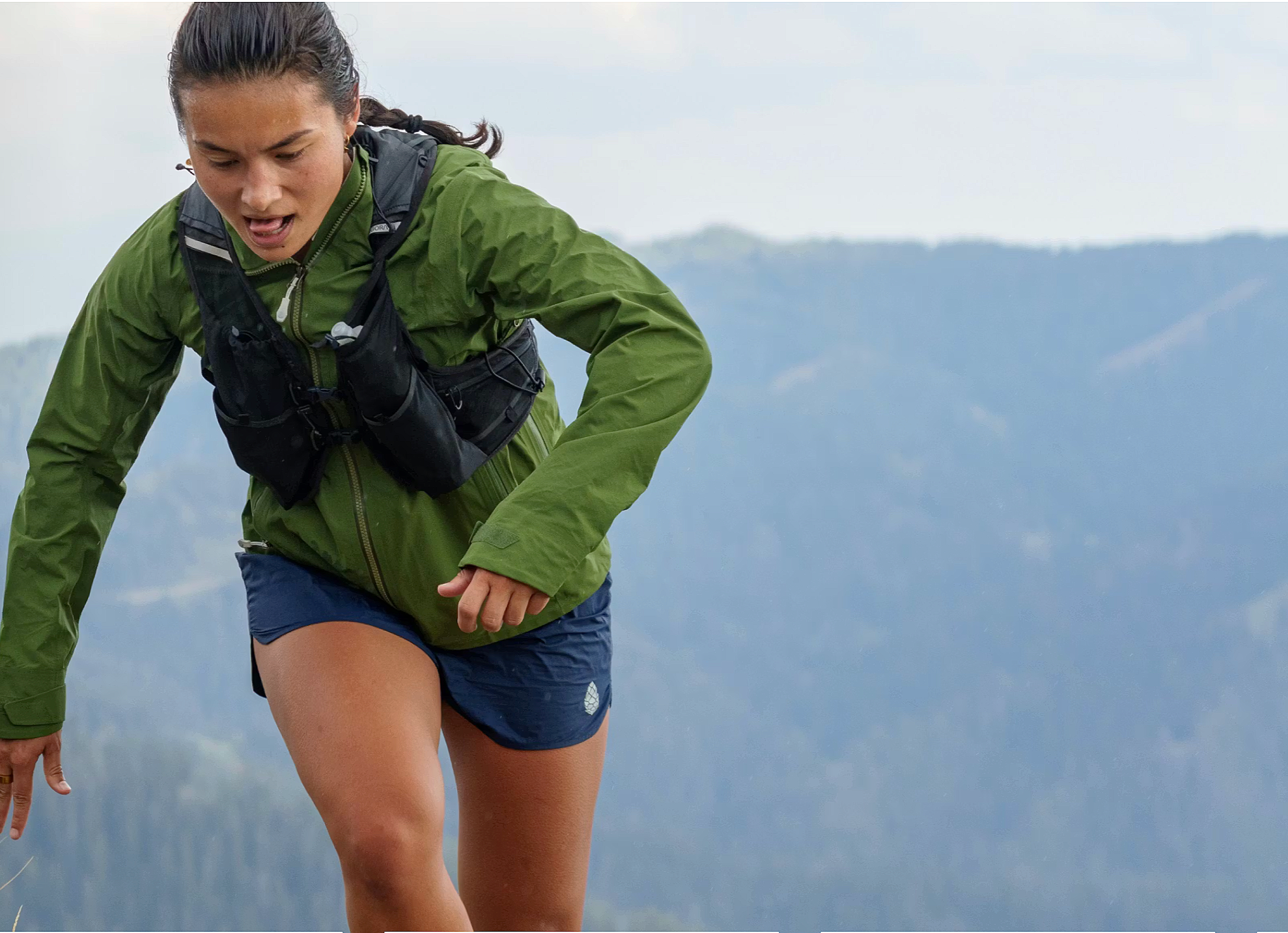 Woman trail running in the mountains wearing a lightweight green jacket and running vest