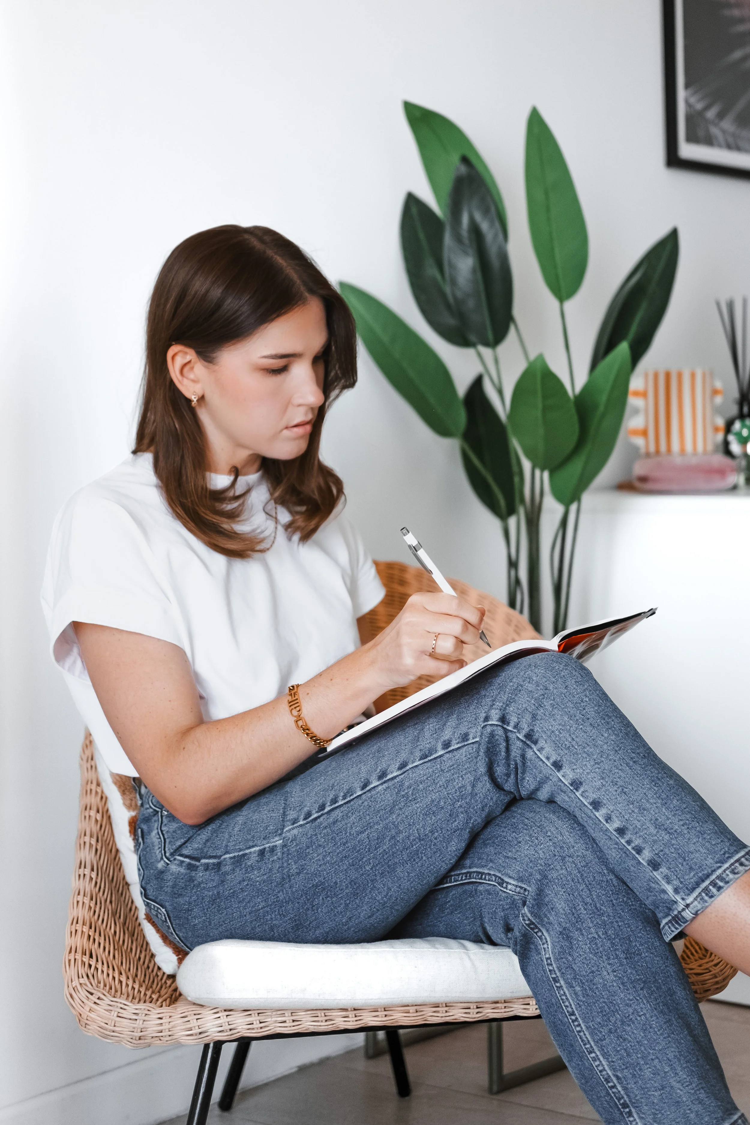 Emily Banks sitting in a cane chair, writing in a notebook beside a plant