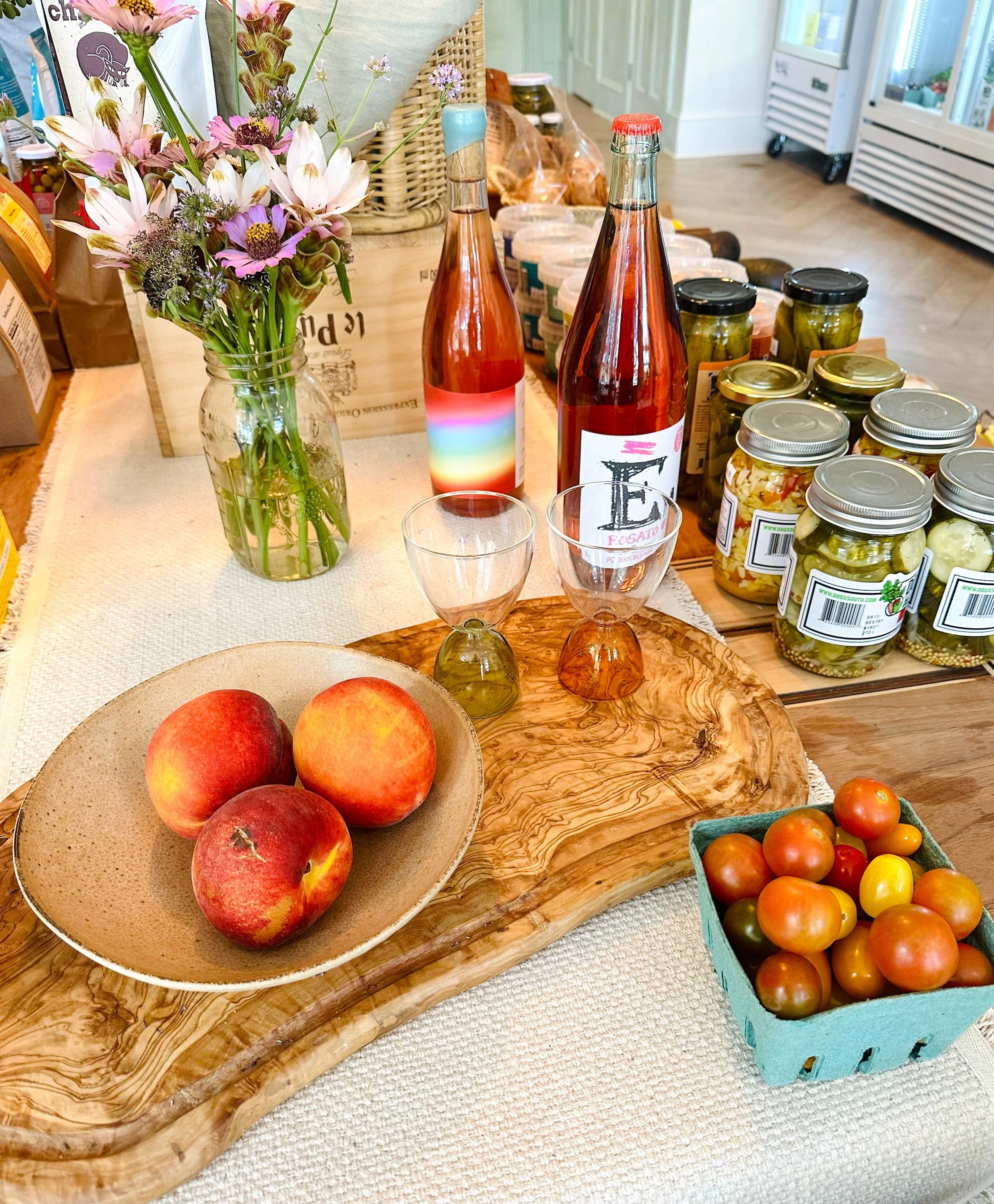 Fresh peaches, heirloom tomatoes, local wine, pickled vegetables, and wildflowers displayed on a rustic wooden board inside Provisions, a neighborhood market in Savannah's Historic District.