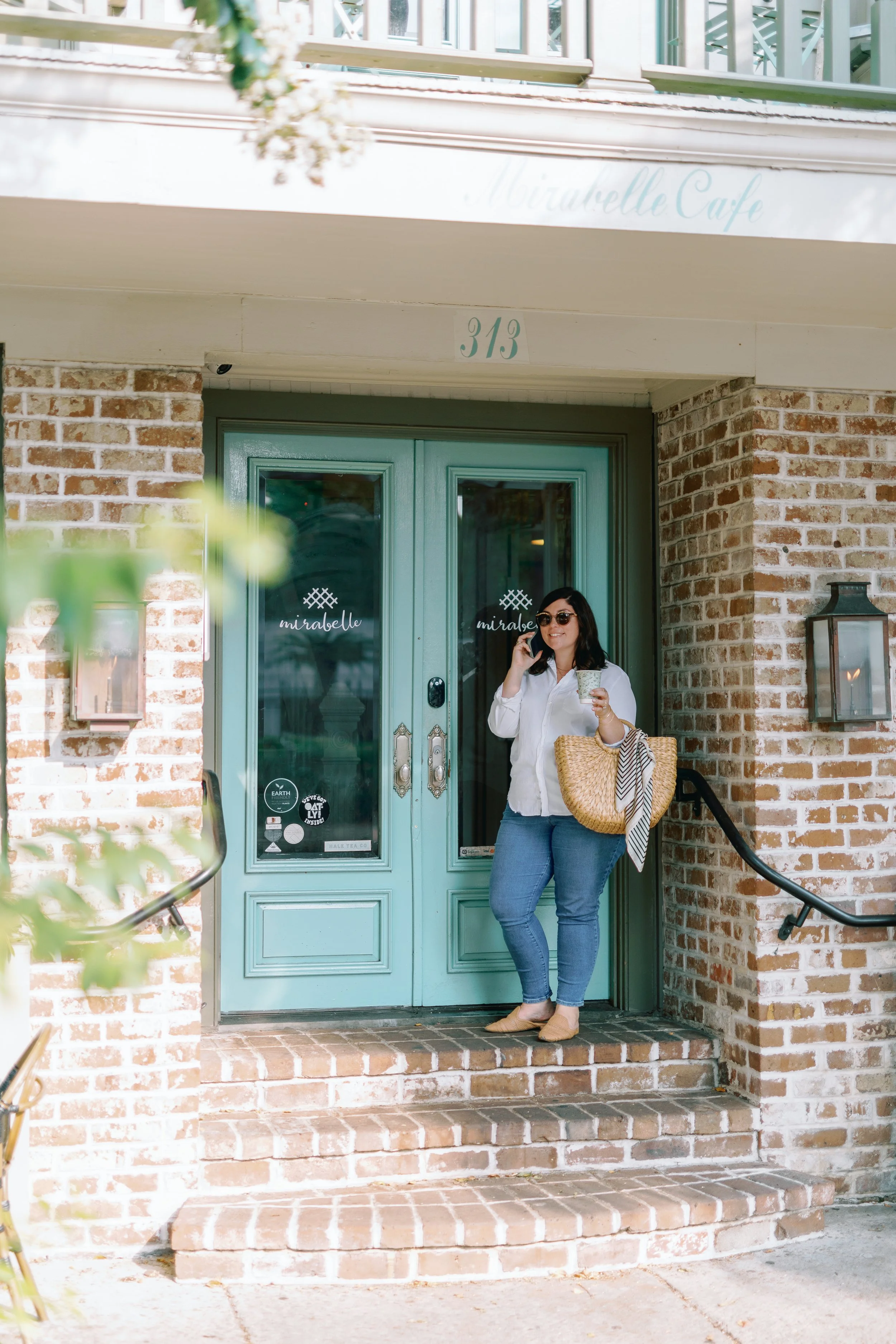 A woman with dark hair and sunglasses, wearing a white blouse and jeans, stands on the steps outside a cafe, holding a drink and talking on her phone.