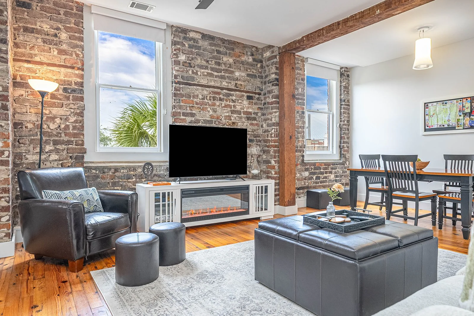 stunnin ghistoric brick wall in confo with electric fireplace and leather furniture and dining room table, exposed wood beams, luxury hosting savannah vacation rental