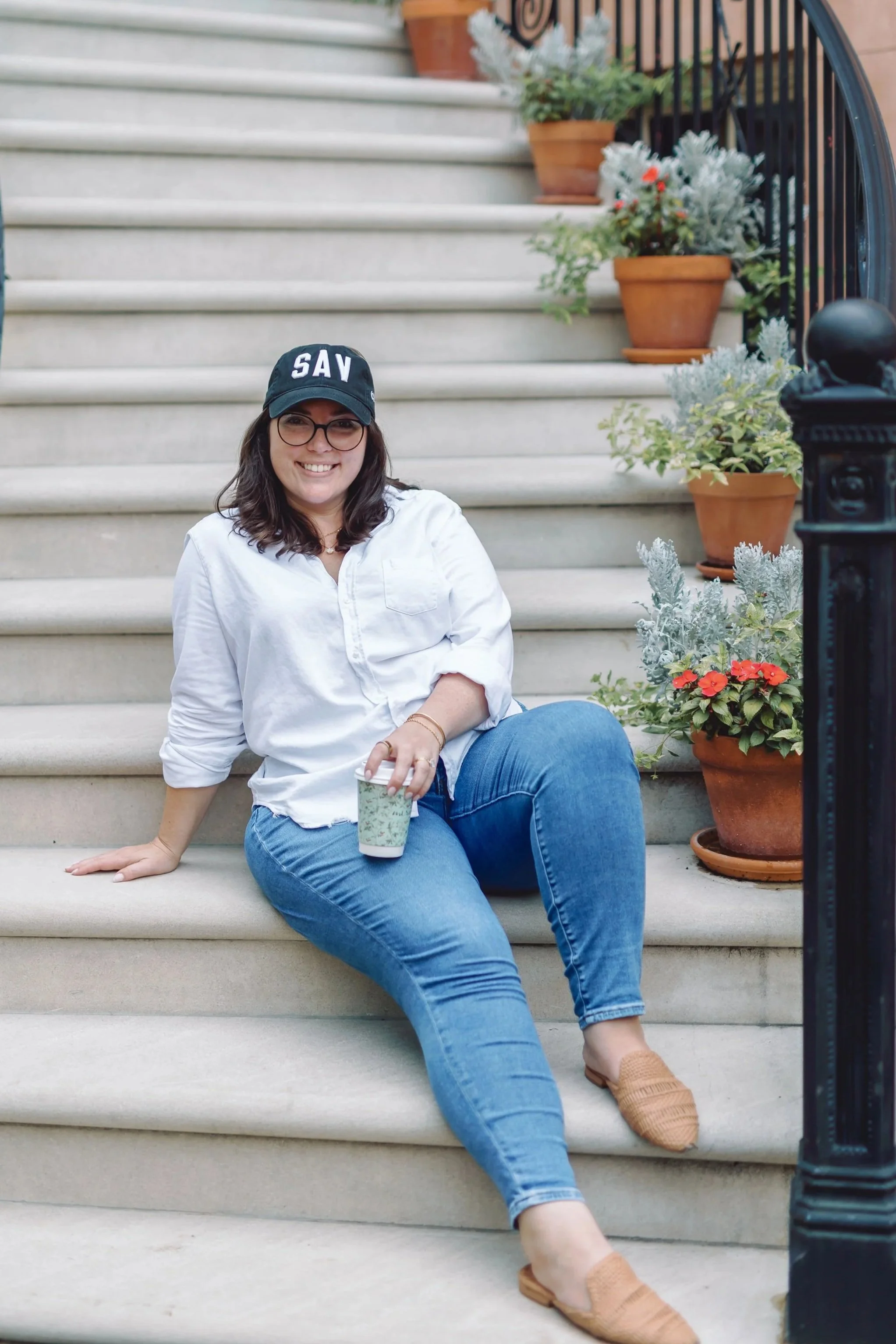 A woman with glasses, wearing a black cap with the word 'SAV', a white shirt, blue jeans, and tan shoes, sitting on outdoor stairs with potted plants along the side.