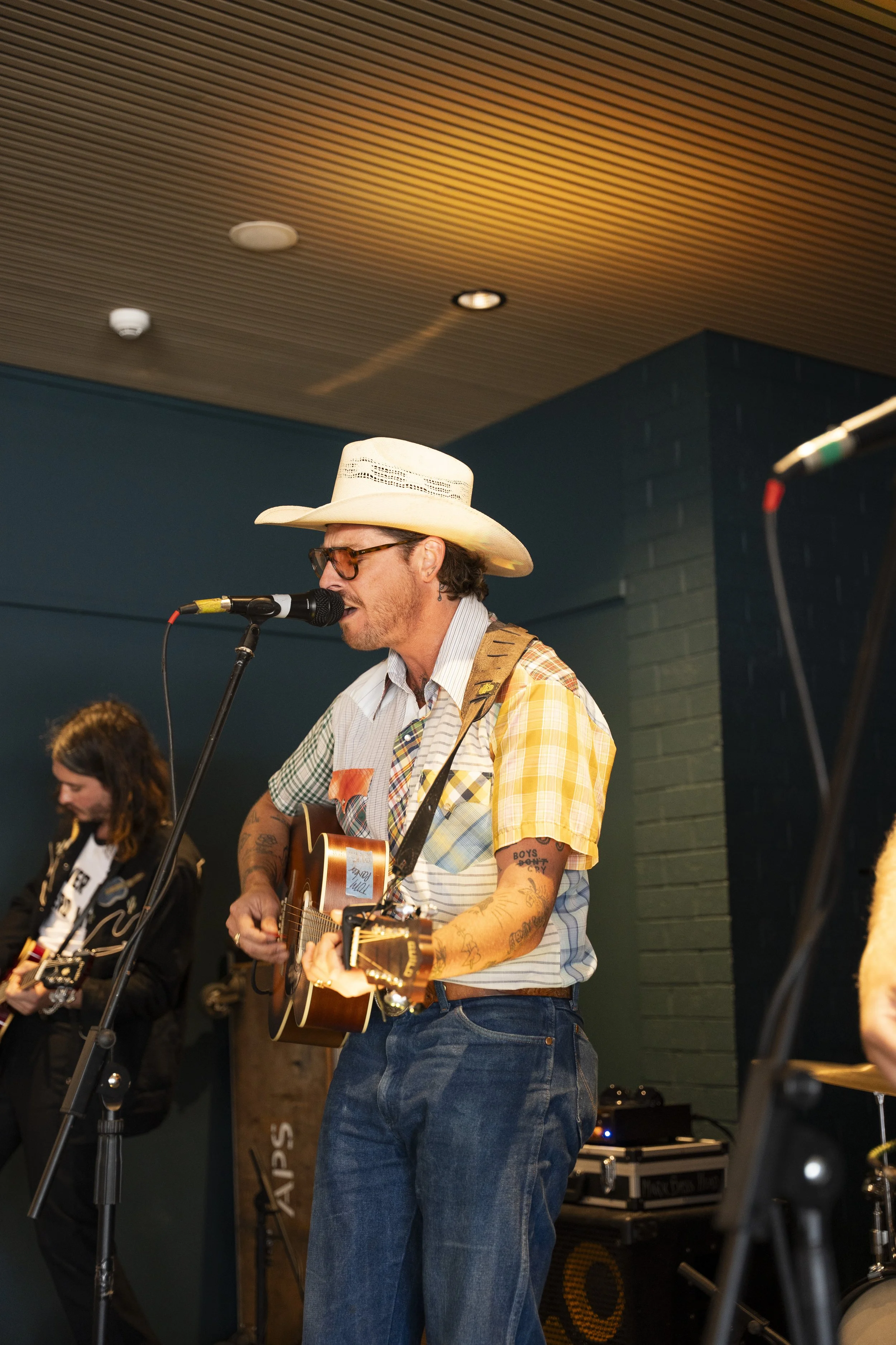 A man wearing a cowboy hat, glasses, and a plaid shirt is playing an acoustic guitar and singing into a microphone during a live performance, with a woman visible in the background.