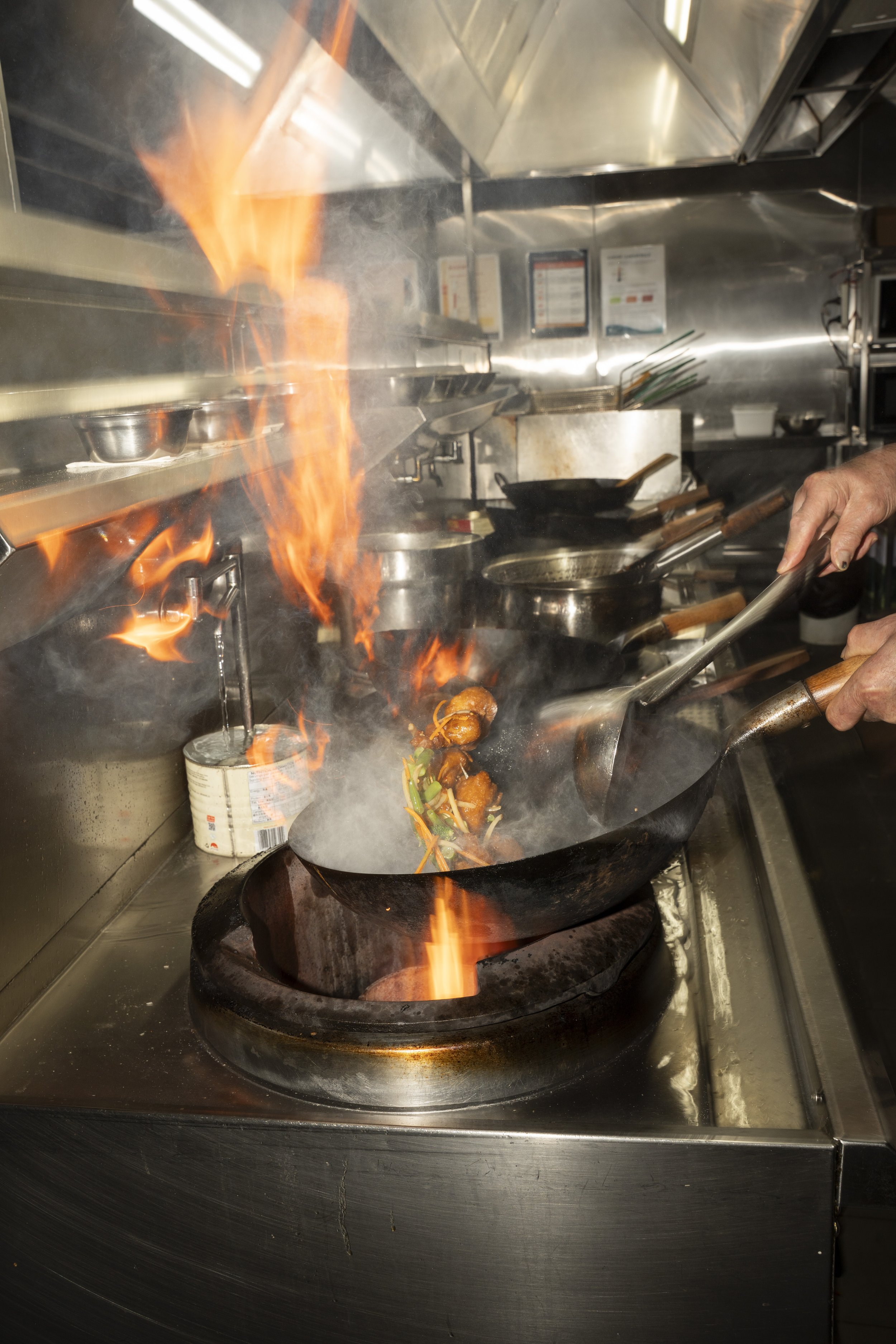 A chef cooking in a kitchen with flames rising from a wok, adding vegetables and sauce.