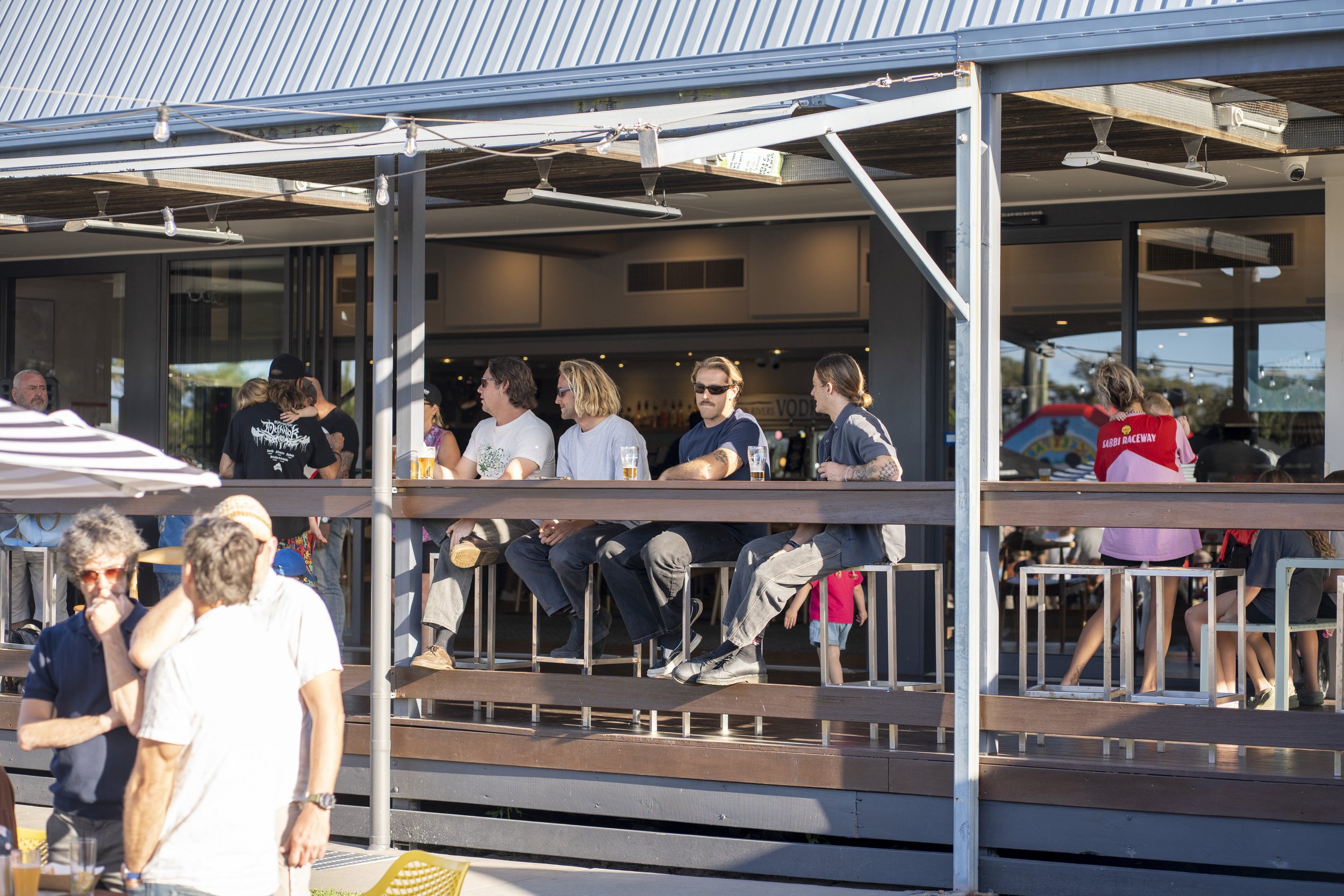 People sitting and standing outside a building with a balcony, some holding drinks, during daylight.