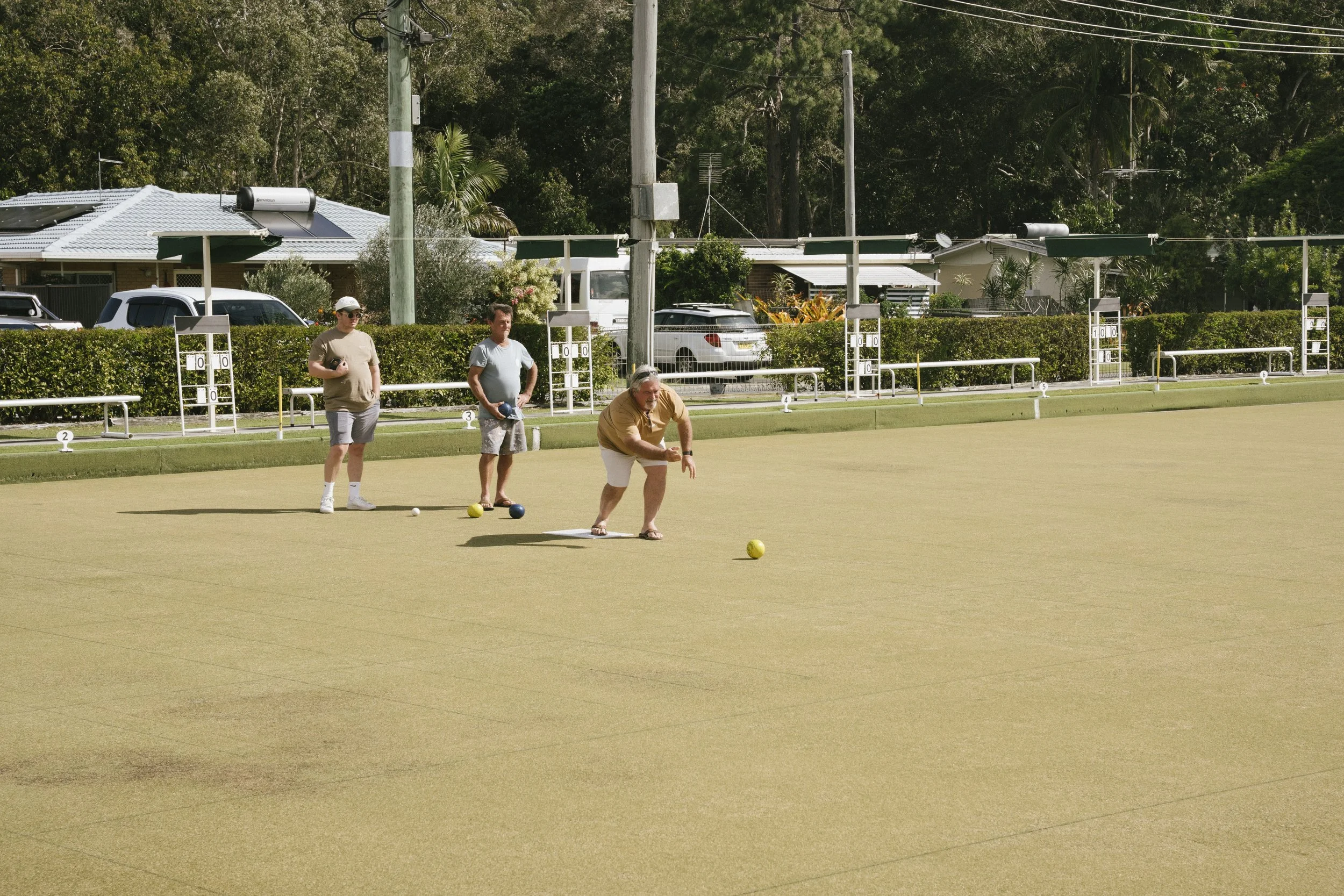 Three men playing lawn bowls on a green field, two of them standing and one crouching to take a shot. In the background, there are houses, parked cars, and tall trees.