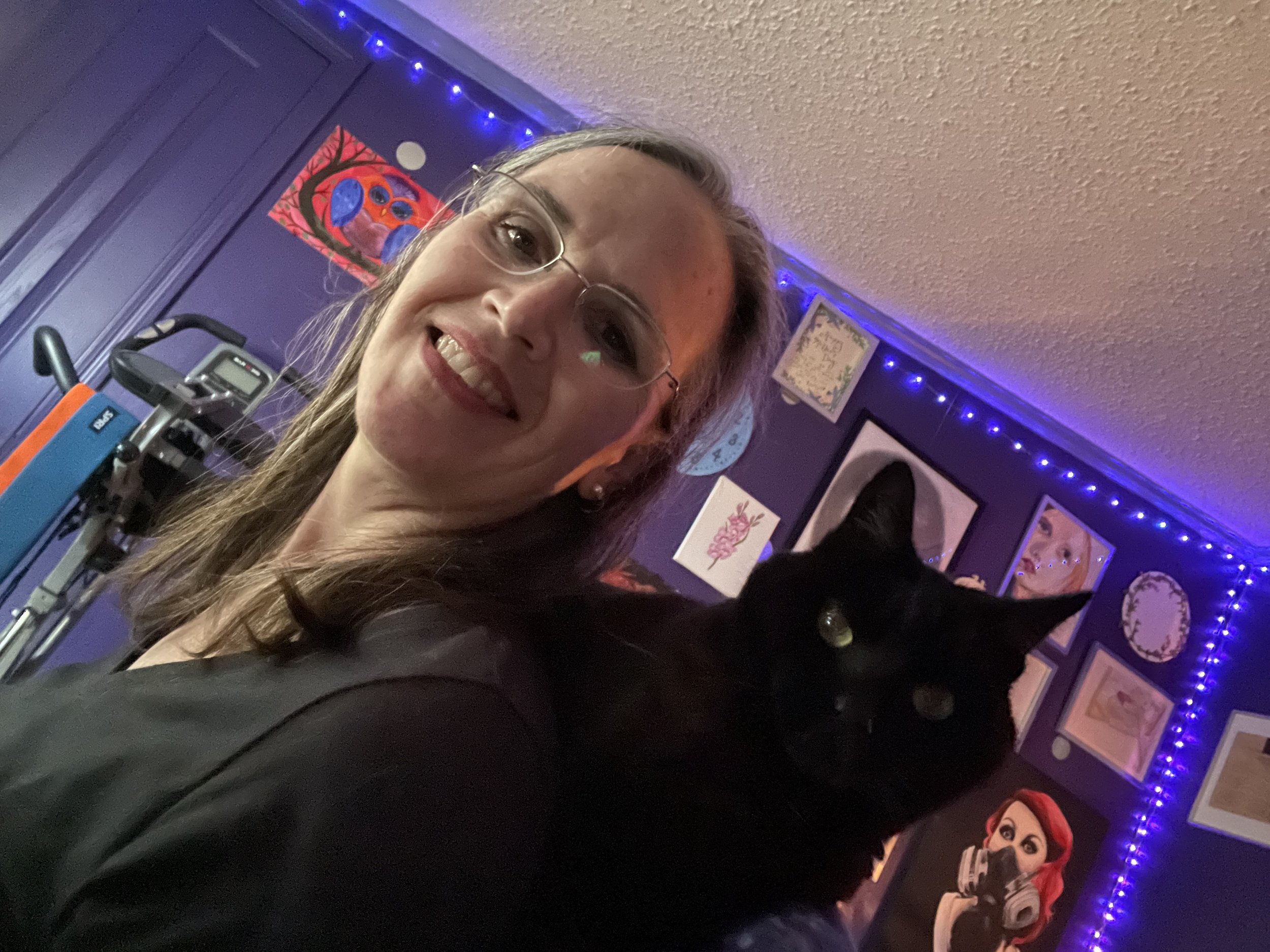 Deelylah, a white woman with shoulder length brown hair with white streaks who also wears glasses, with her furry co-worker, Puck, a black cat, who sits on her shoulders.