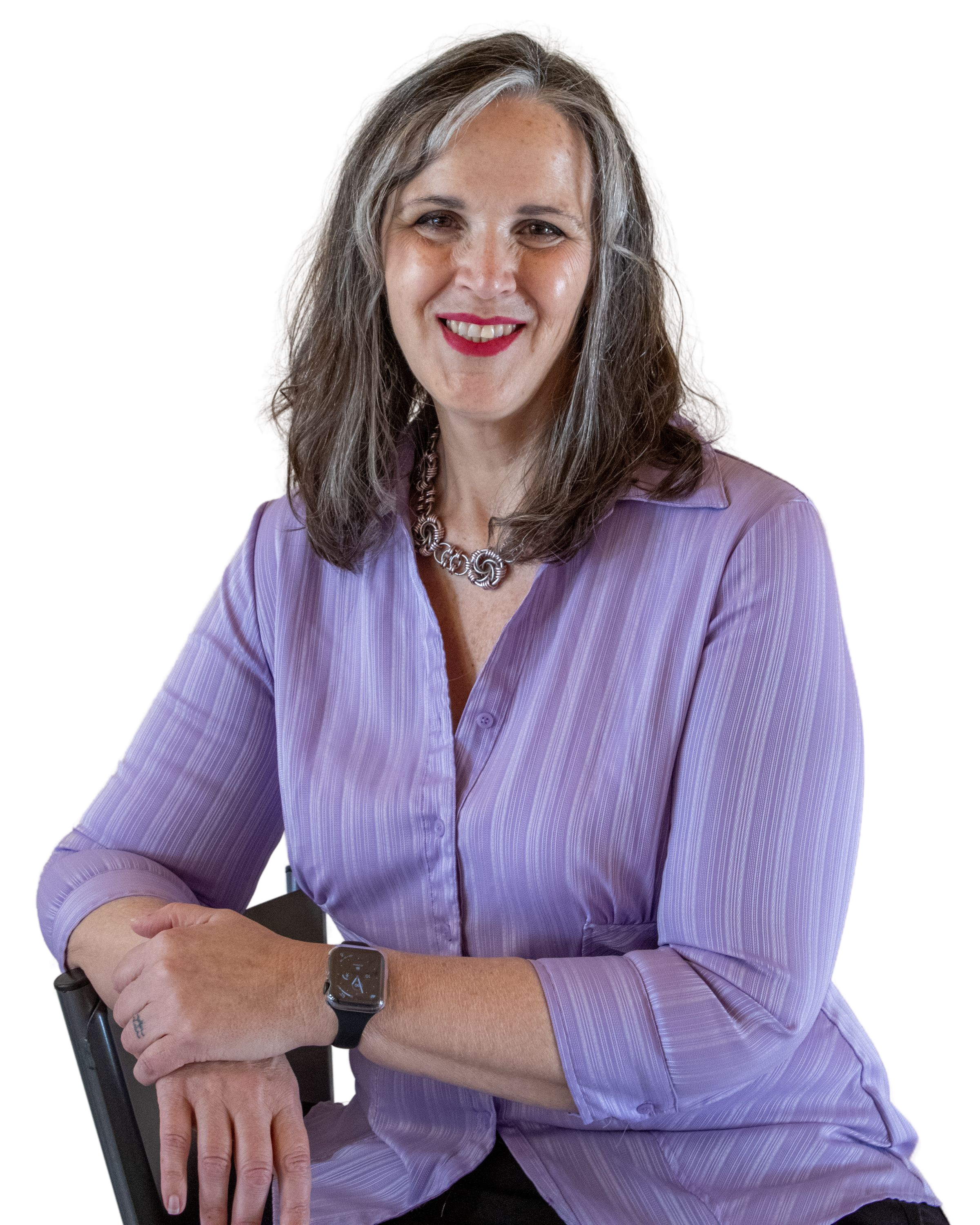 A woman with long gray hair wearing a lavender button-up shirt, a silver necklace, and a black wristwatch is seated, smiling, and resting her illuminated smartwatch on her left wrist.