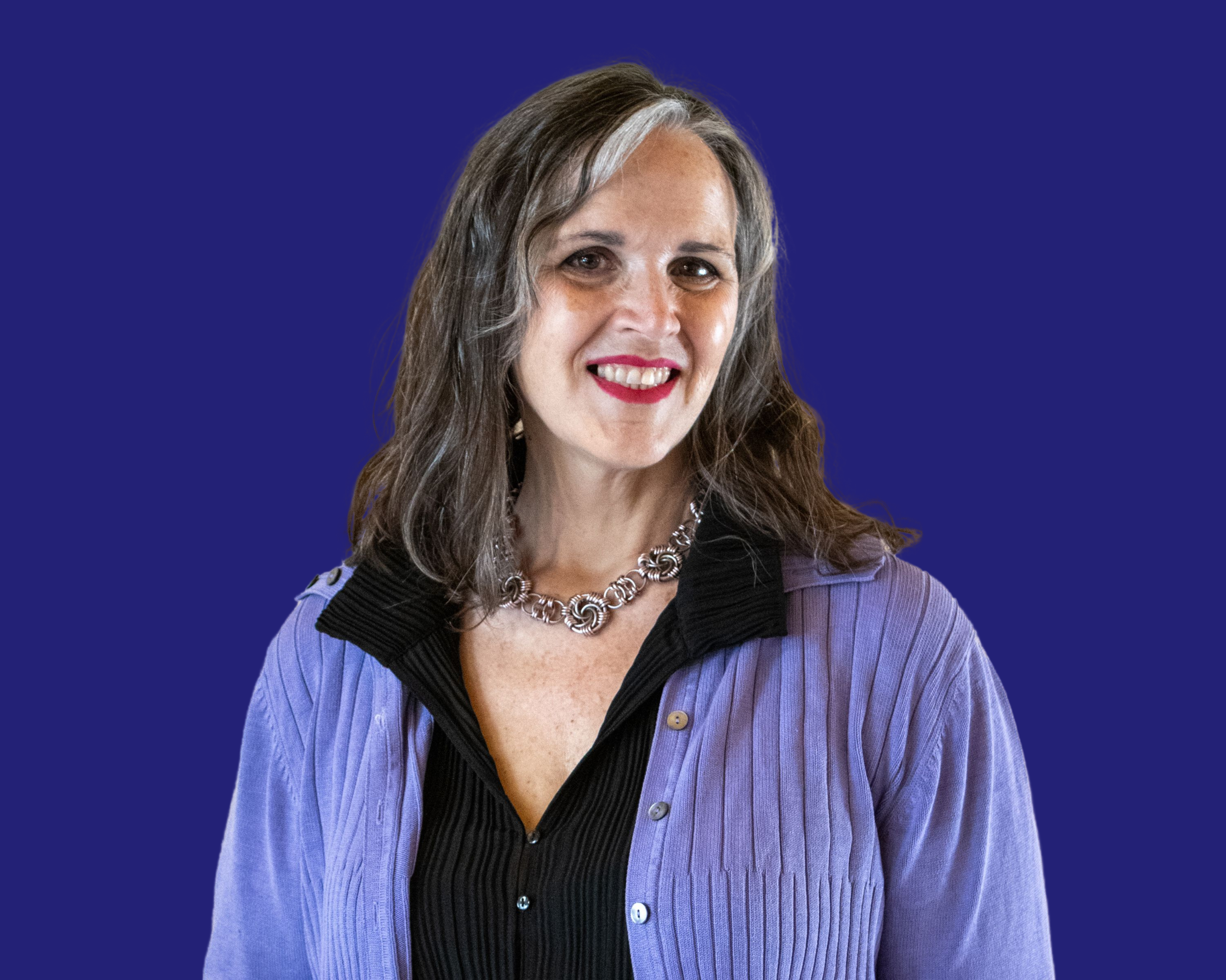 A woman with brown hair and gray streaks, smiling, wearing a purple blazer over a black shirt, and a silver necklace, against a solid blue background.