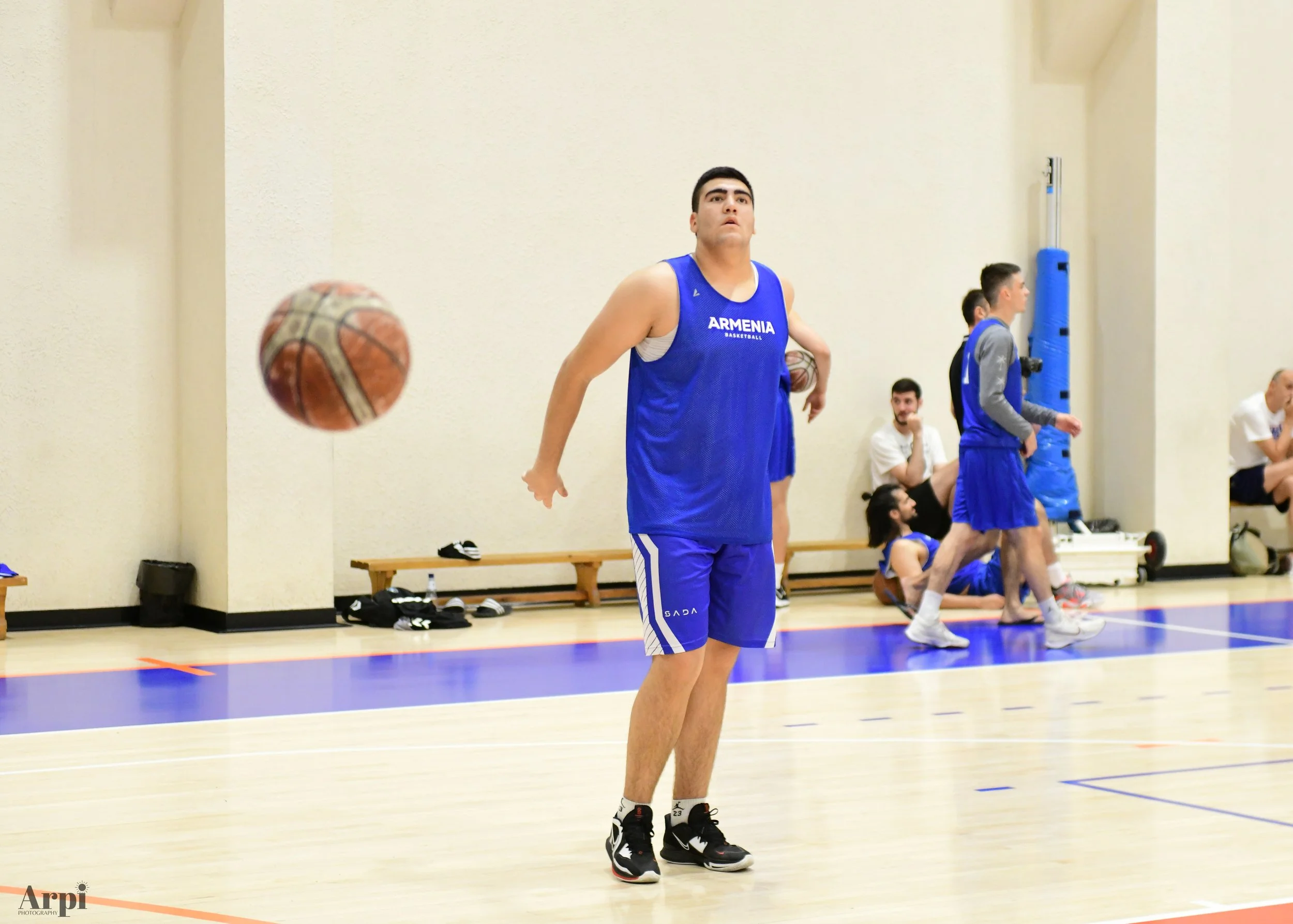 Basketball player in blue "Armenia" jersey on indoor court during training, with basketball in motion and other players in background.