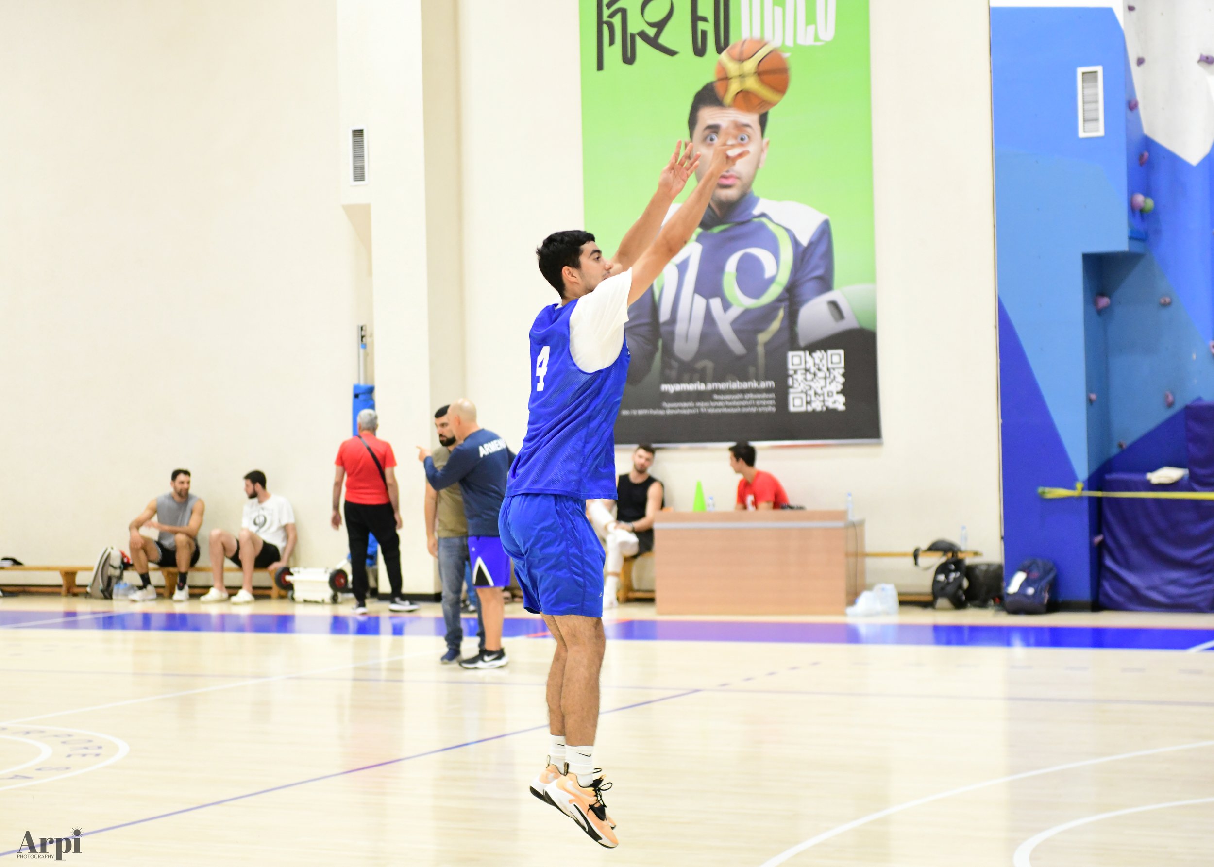 Basketball player in blue jersey shooting a jump shot in an indoor gym, with people and a poster in the background.