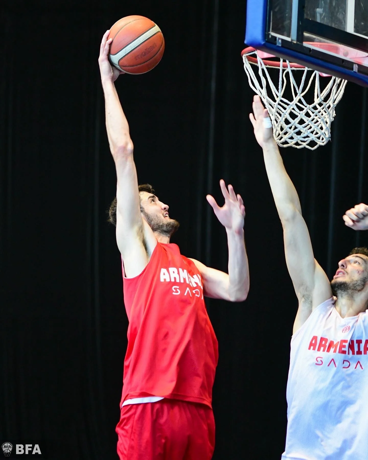 Two basketball players in Armenia jerseys during a game, one in a red uniform jumping to make a shot with the ball near the hoop, and the other in a white uniform attempting to block.