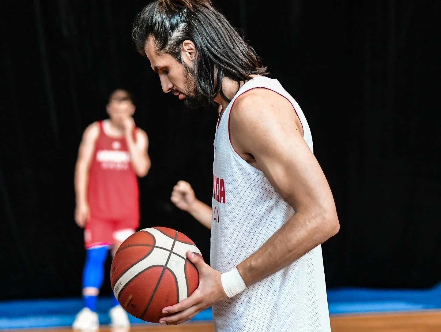 Basketball player in white jersey holding a basketball, with another player in the background wearing red jersey, on an indoor court.